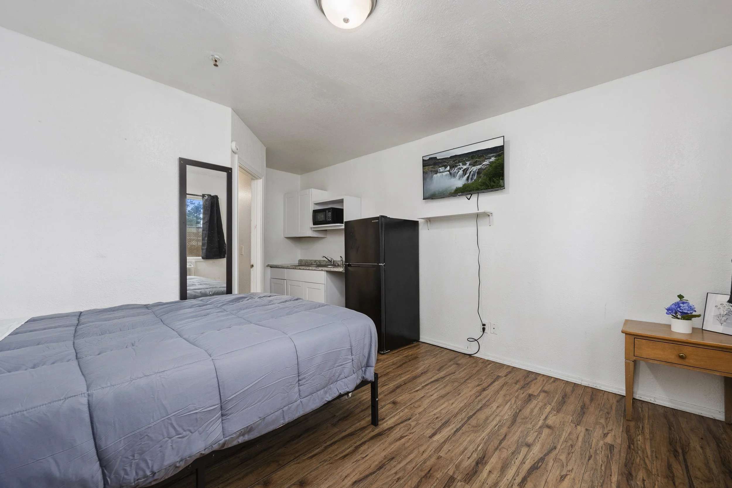 A bedroom with a bed, a dresser with a flowerpot, a kitchen area with cabinets, a microwave, a refrigerator, a mounted flat-screen TV showing a waterfall, and a window with a black curtain.