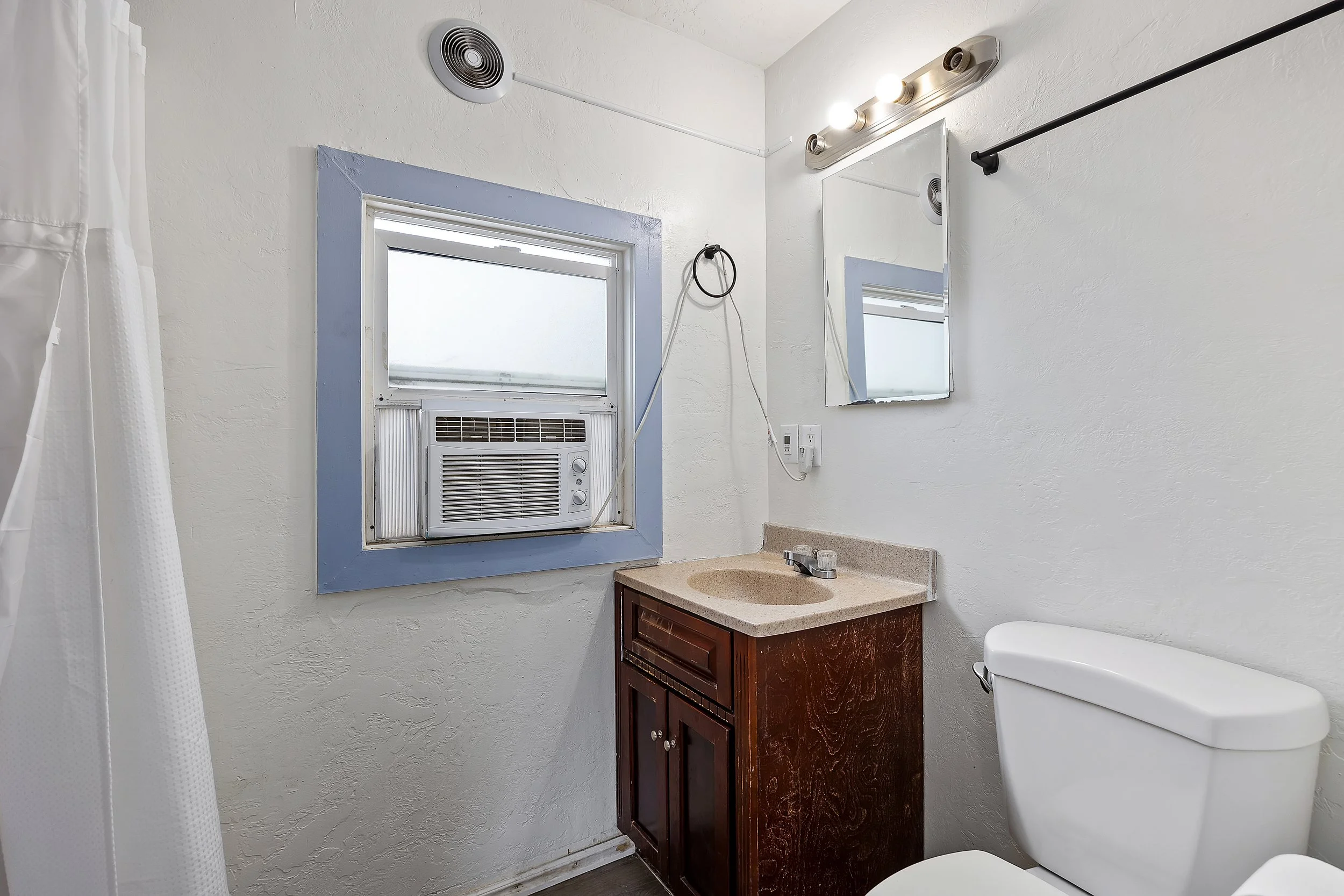 Bathroom with a window air conditioning unit, a mirror, a vanity with a brown wooden cabinet, a beige sink, a white toilet, and a white shower curtain. The walls are white with a blue trim around the window, and there is an overhead light fixture wit