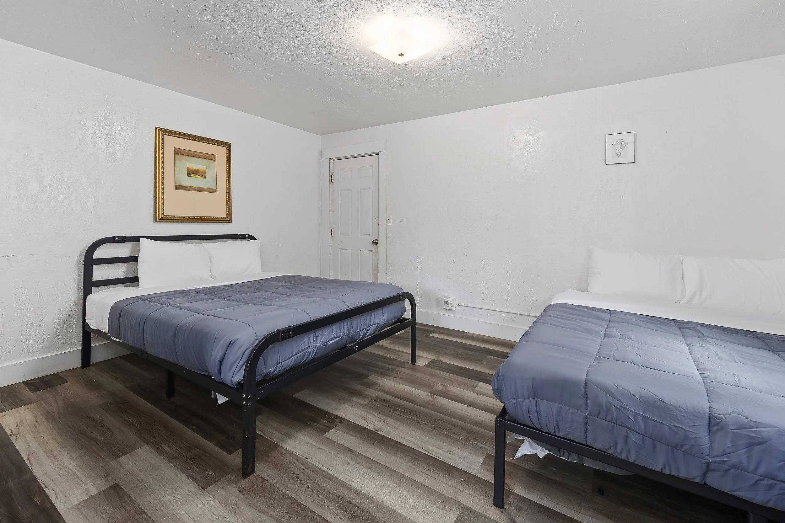 Two beds with grey bedding and white pillows in a room with wood flooring, white textured walls, a white door, and framed pictures hung on the walls.