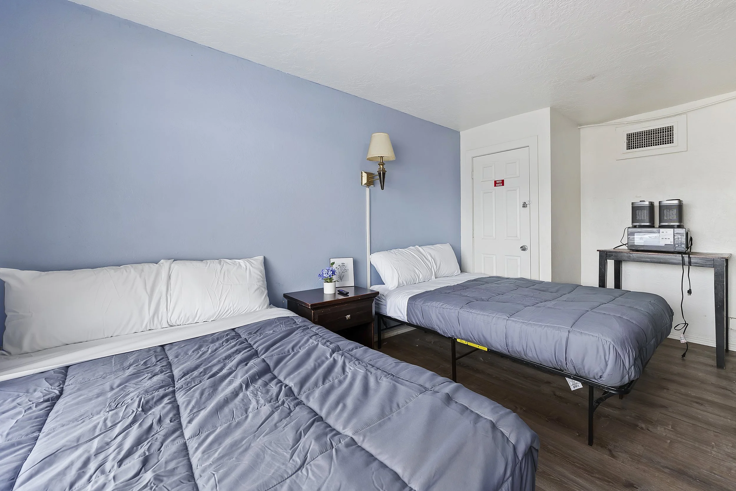 A hotel room with two beds covered with gray quilts, a nightstand with a small flower pot and a remote control, a wall-mounted lamp, a door with a red 'Water Heater' sign, and a small table holding a microwave, a coffee maker, and two water heaters i