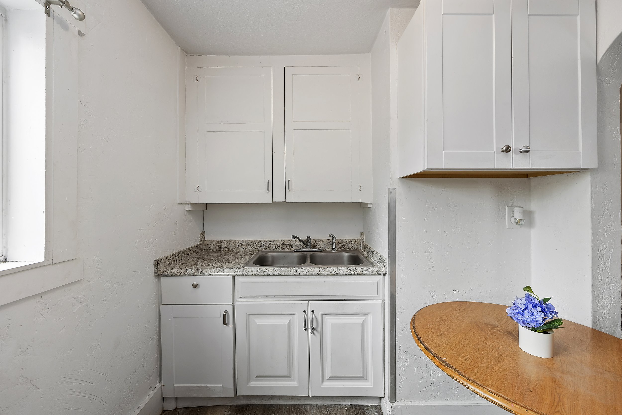 A small kitchen corner with white cabinets, a granite countertop, a double sink, some plant decor on a wooden table, and a window on the left wall.