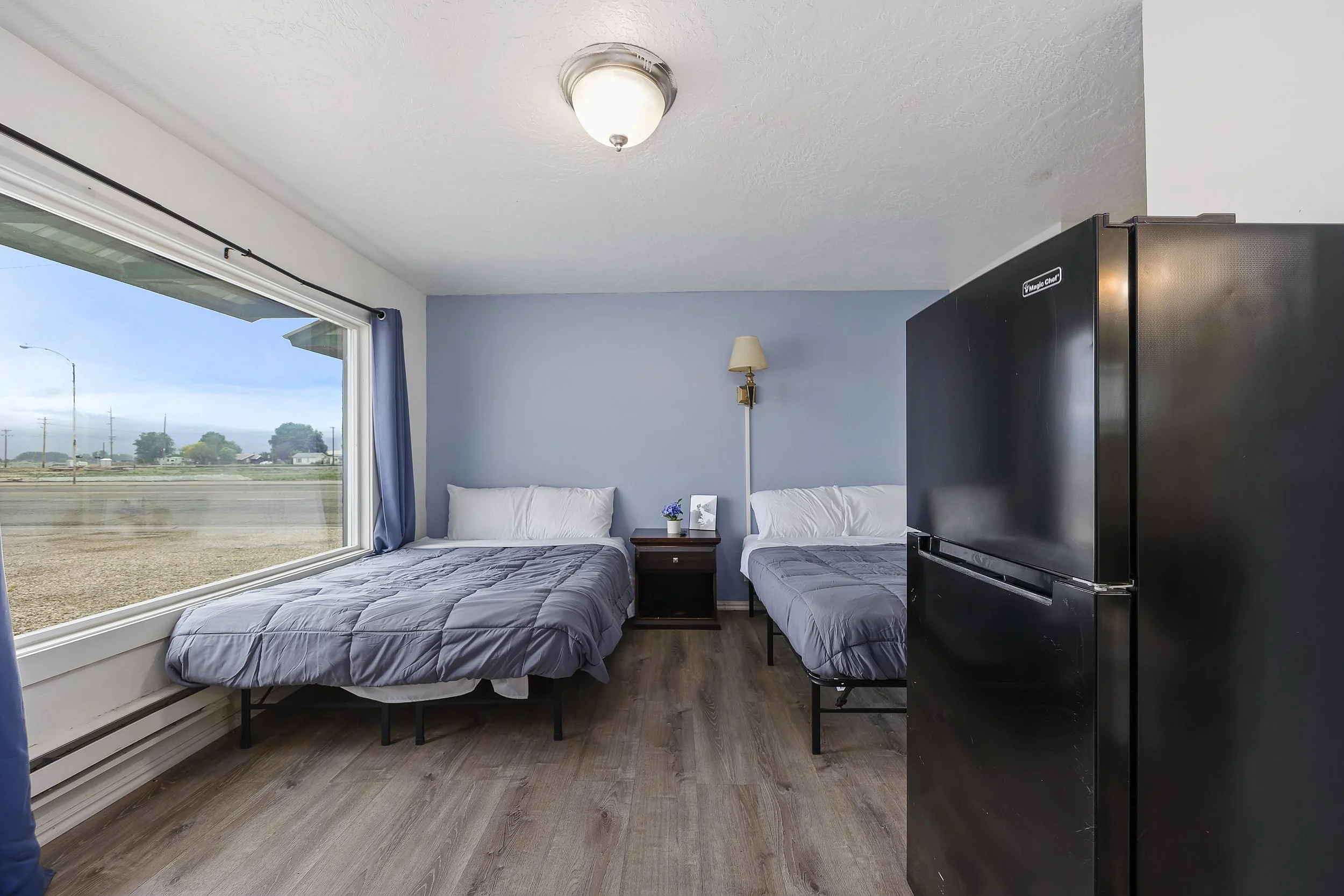 A hotel room with two beds, a window, a nightstand, a wall lamp, and a refrigerator.
