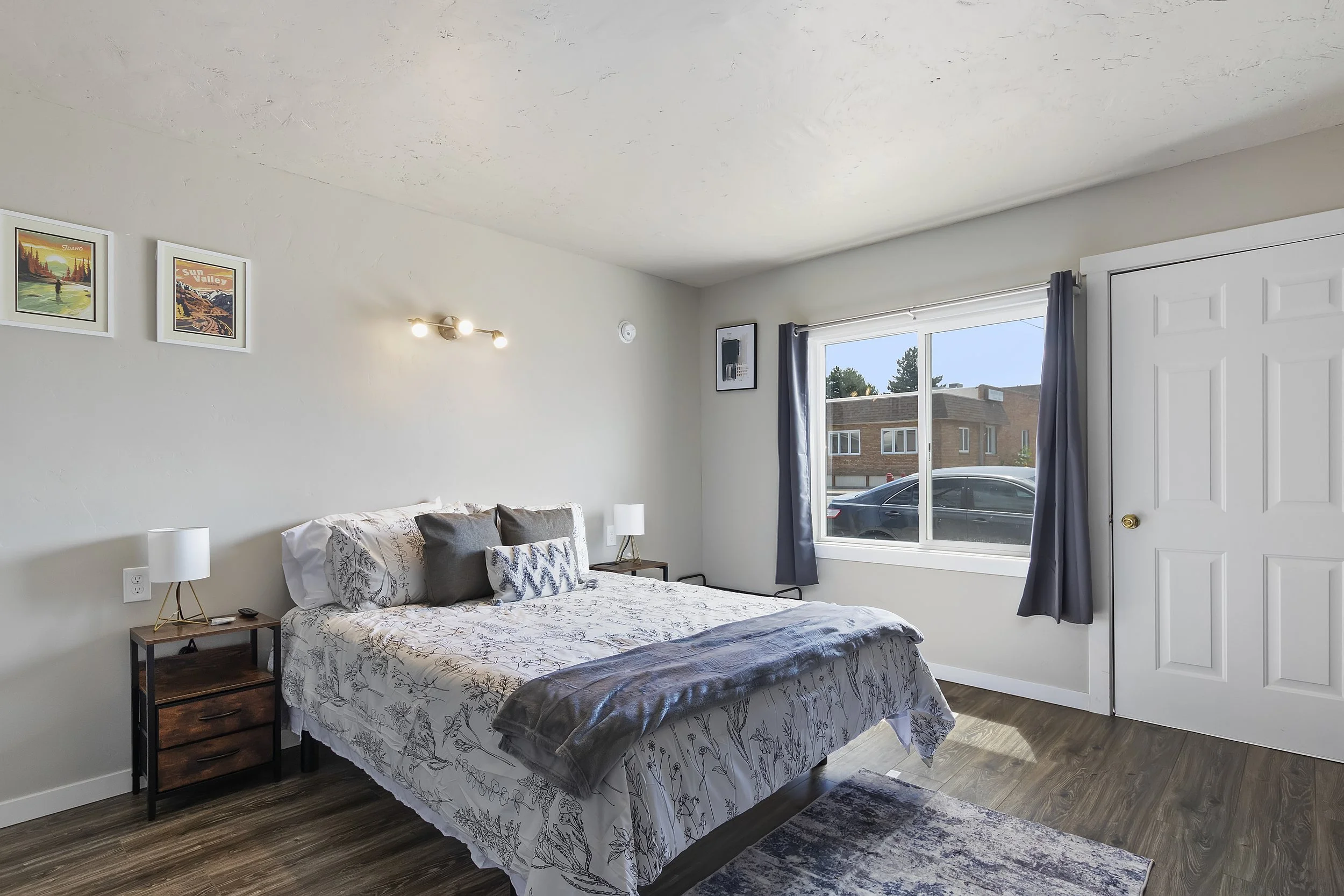 A bedroom with a bed covered in floral-patterned bedding, two pillows, and a brown velvet blanket. There are two nightstands with white lamps, a window with a view of neighboring buildings, and framed artwork on the wall.