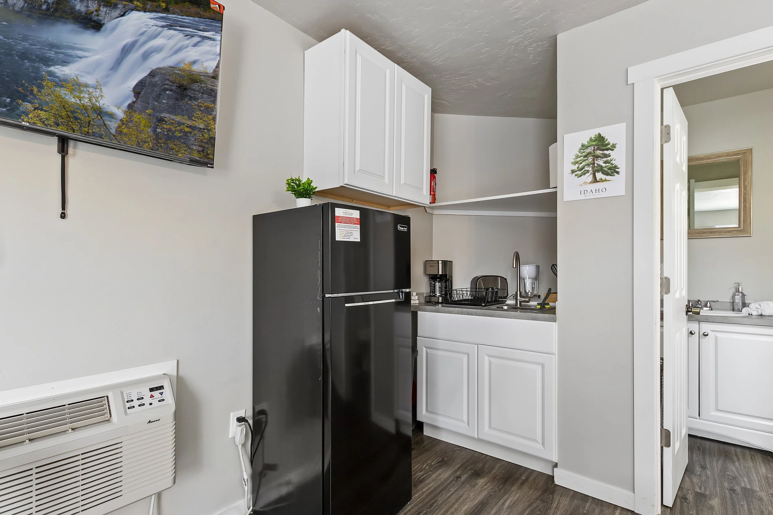 A small kitchen with a black refrigerator, white upper cabinets, a sink, and countertop appliances, with a wall art poster of a tree labeled Idaho, and a view into a room with a mirror.