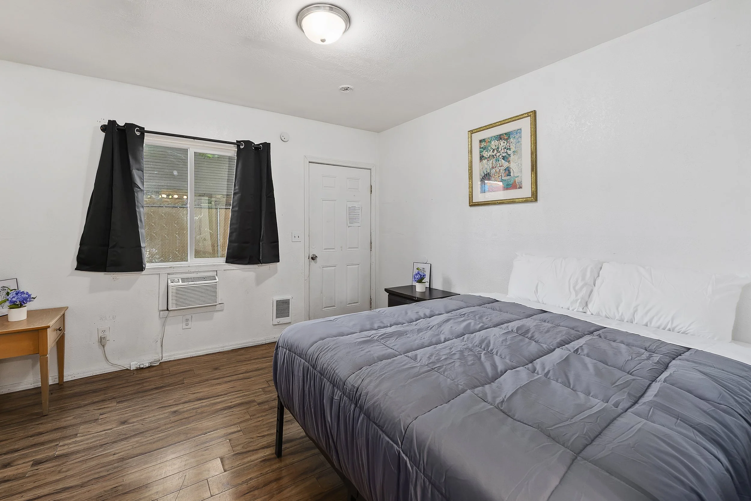 A bedroom with a bed covered in a gray comforter, white pillows, a nightstand with a small framed picture and a potted plant, a window with black curtains, a door, hardwood flooring, a framed floral painting on the wall, and a ceiling light.