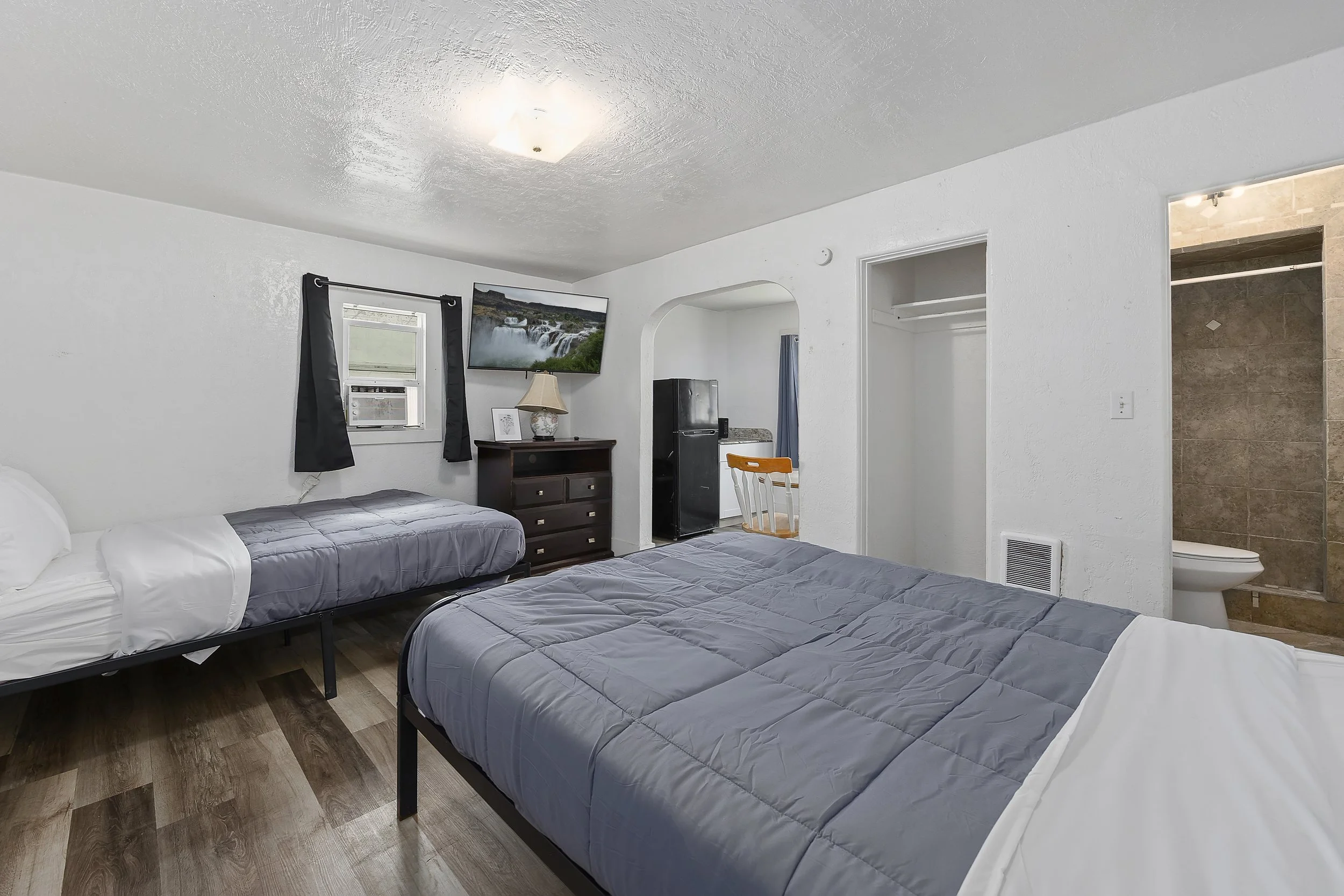 A bedroom with two beds covered in gray comforters and white pillows, a wooden dresser with a table lamp, a small refrigerator, a TV mounted on the wall showing a waterfall scene, a window with black curtains, and a view of an open bathroom area with