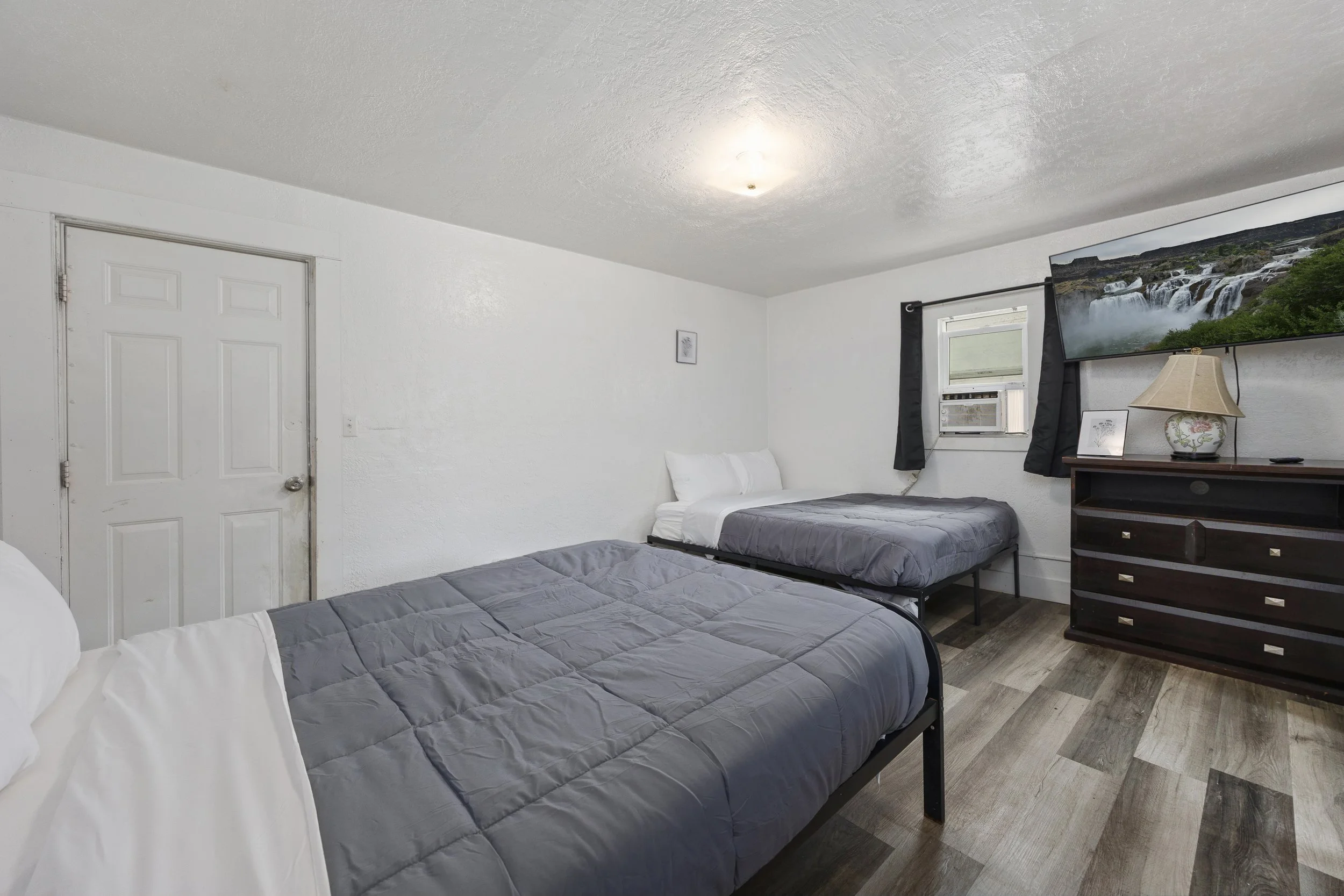 A bedroom with white walls, two beds with gray comforters, a dresser, a lamp, and a window with black curtains showing a waterfall scene on the TV.