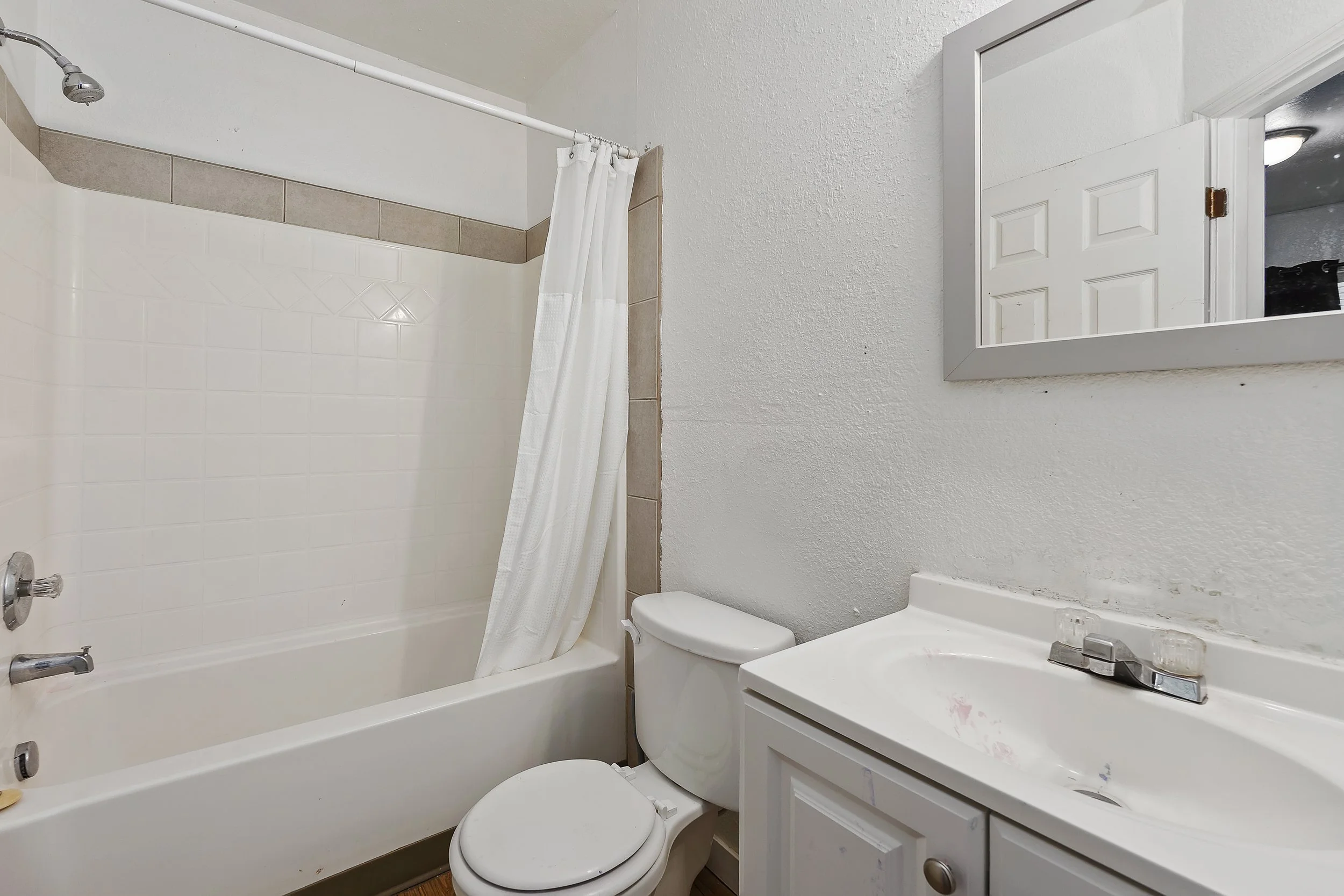 A small bathroom with a shower/tub combination, a white toilet, and a white vanity with a sink. There is a mirror above the vanity and a small cabinet on the wall next to the mirror. The shower has a shower curtain and a silver showerhead. The walls 