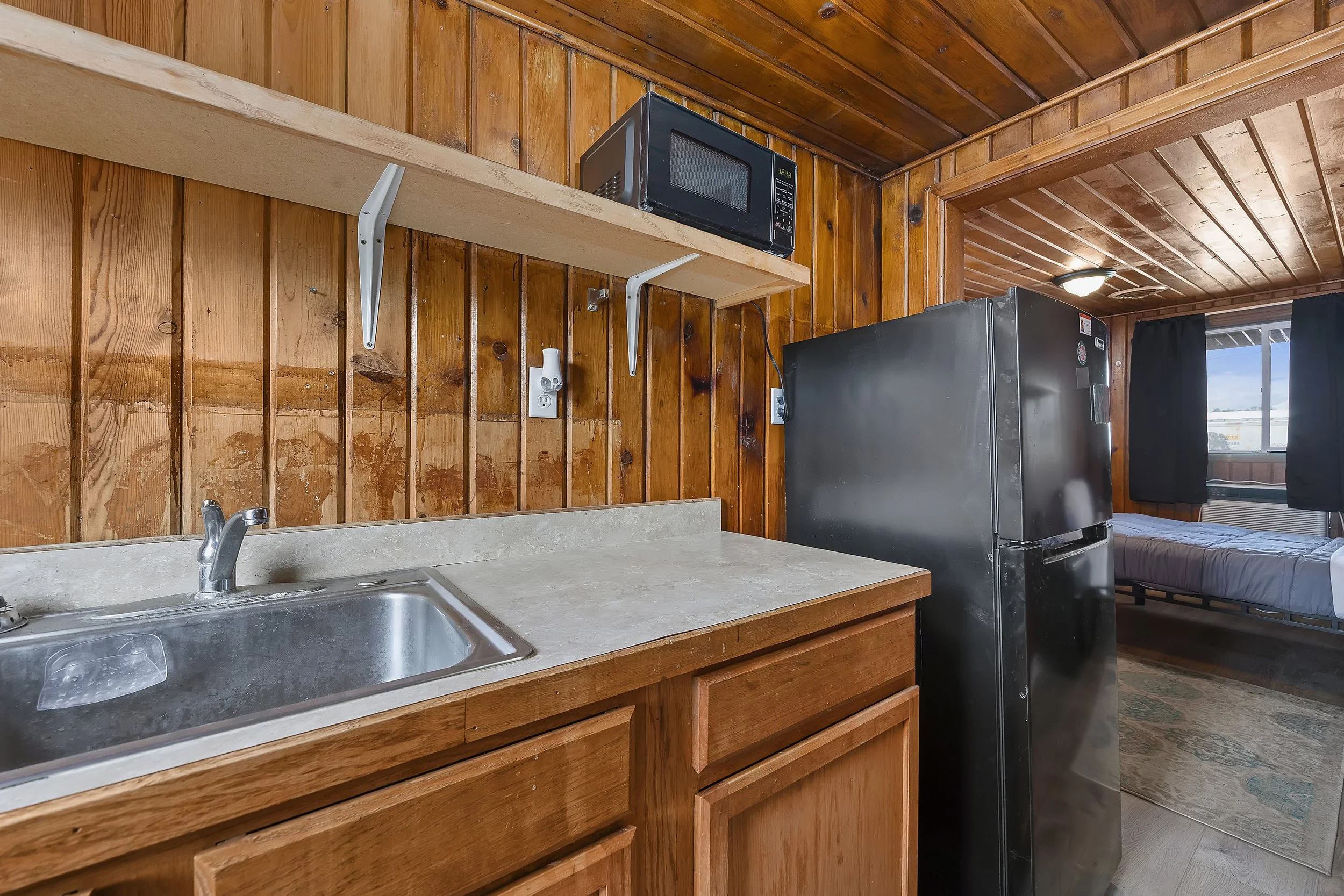 Wooden kitchen with a stainless steel sink, countertop, microwave on a wooden shelf, and a refrigerator. A window with black curtains is visible in the background.