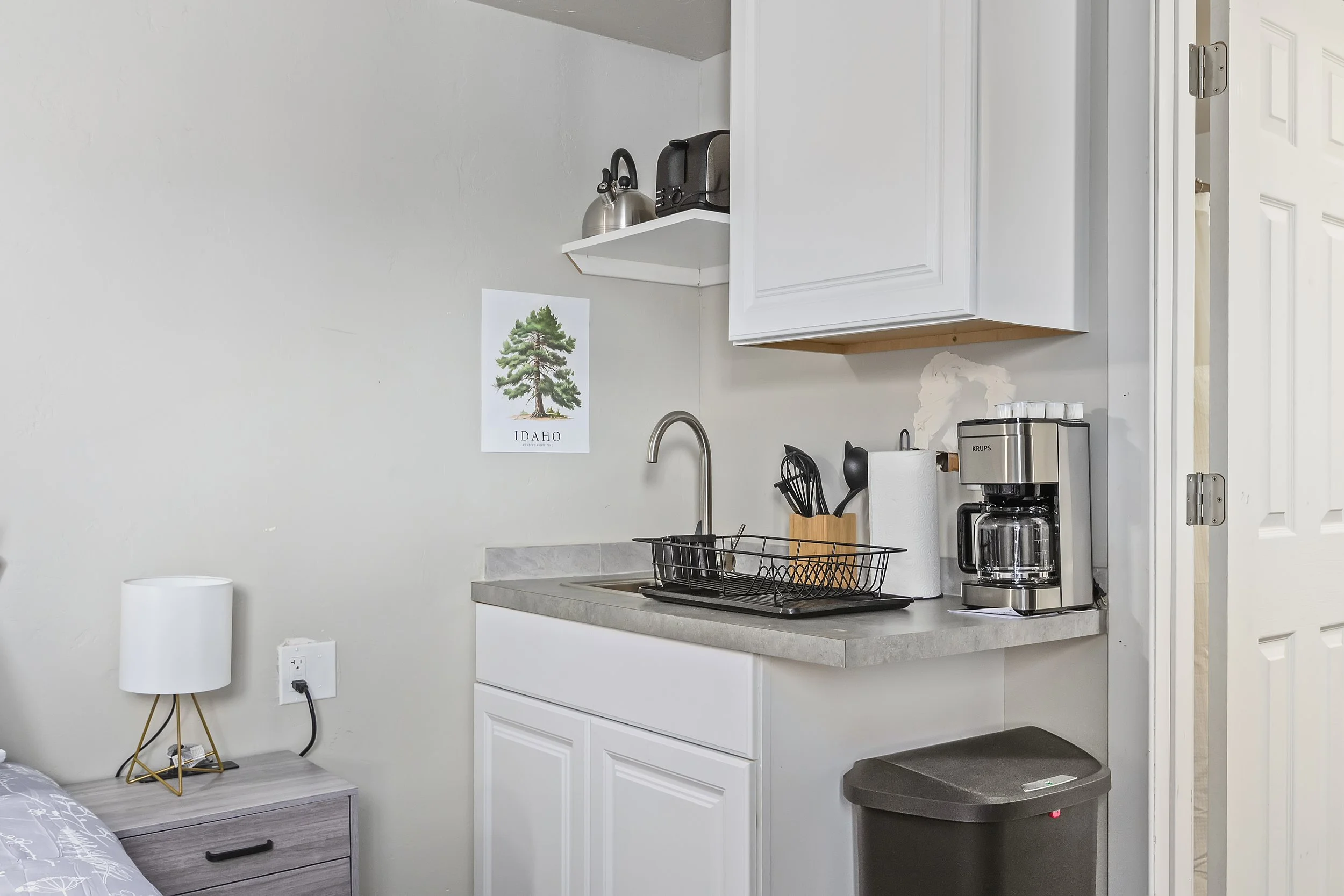 A small kitchen with white cabinets, a countertop, and a sink. On the wall is a poster of a tree with 'IDAHO' written underneath. Items include a toaster, kettle, coffee maker, paper towel roll, utensil holder, and a laundry bin.