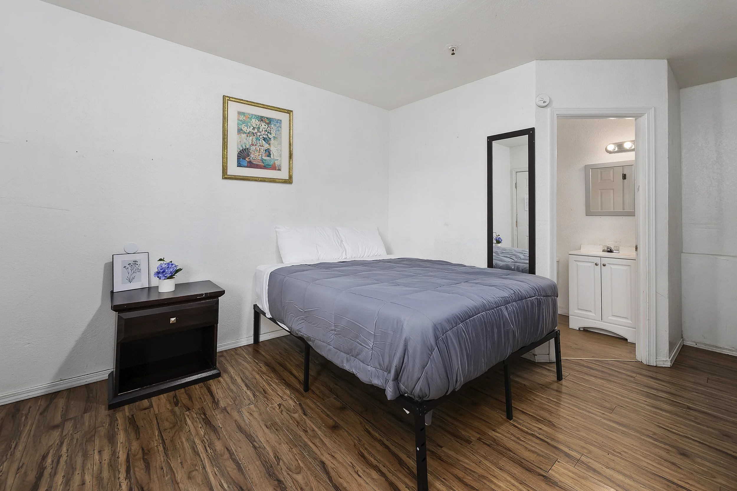 A bedroom with a bed with a gray quilt, a small black nightstand with a framed picture, a small flower in a white pot, and a framed picture hanging above the bed. There is a white bathroom with a mirror and light fixture visible through an open door.
