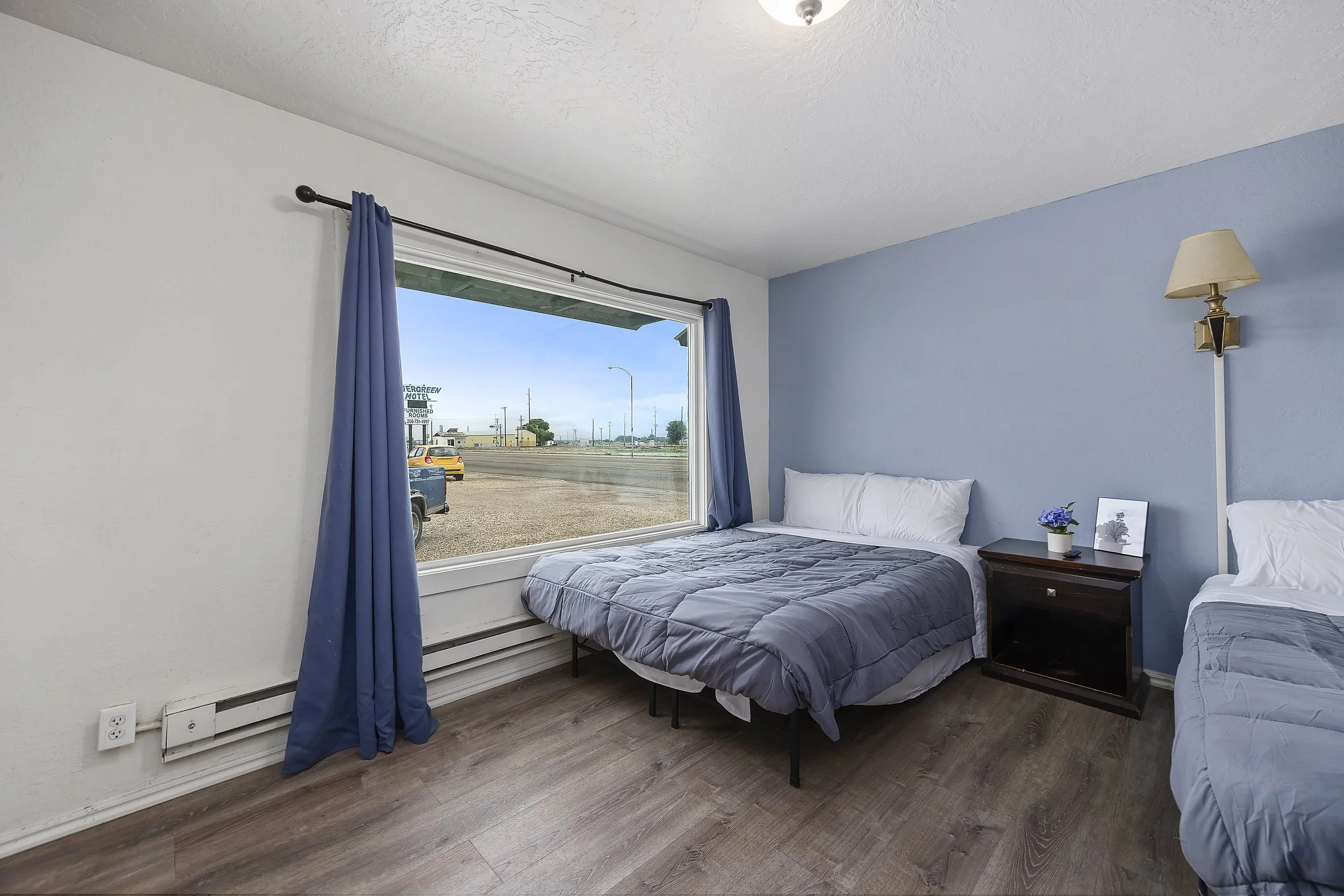 A bedroom with two twin beds, one with gray bedding, a nightstand with a flowerpot, and a window with blue curtains showing a street view outside.