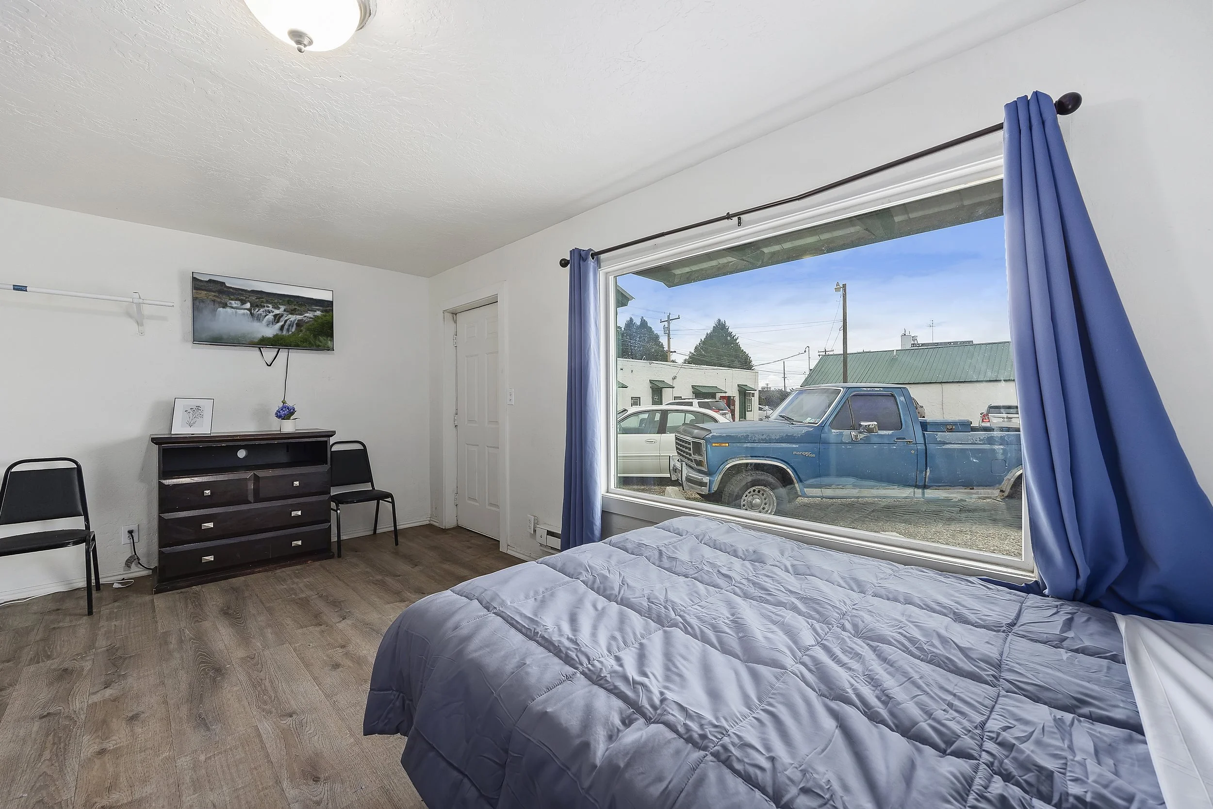 A bedroom with a large window showing a blue truck parked outside, with neighboring buildings and power lines visible. The room has a bed with a gray comforter, a small dark wooden dresser, three black chairs, a wall-mounted TV, blue curtains, and a 