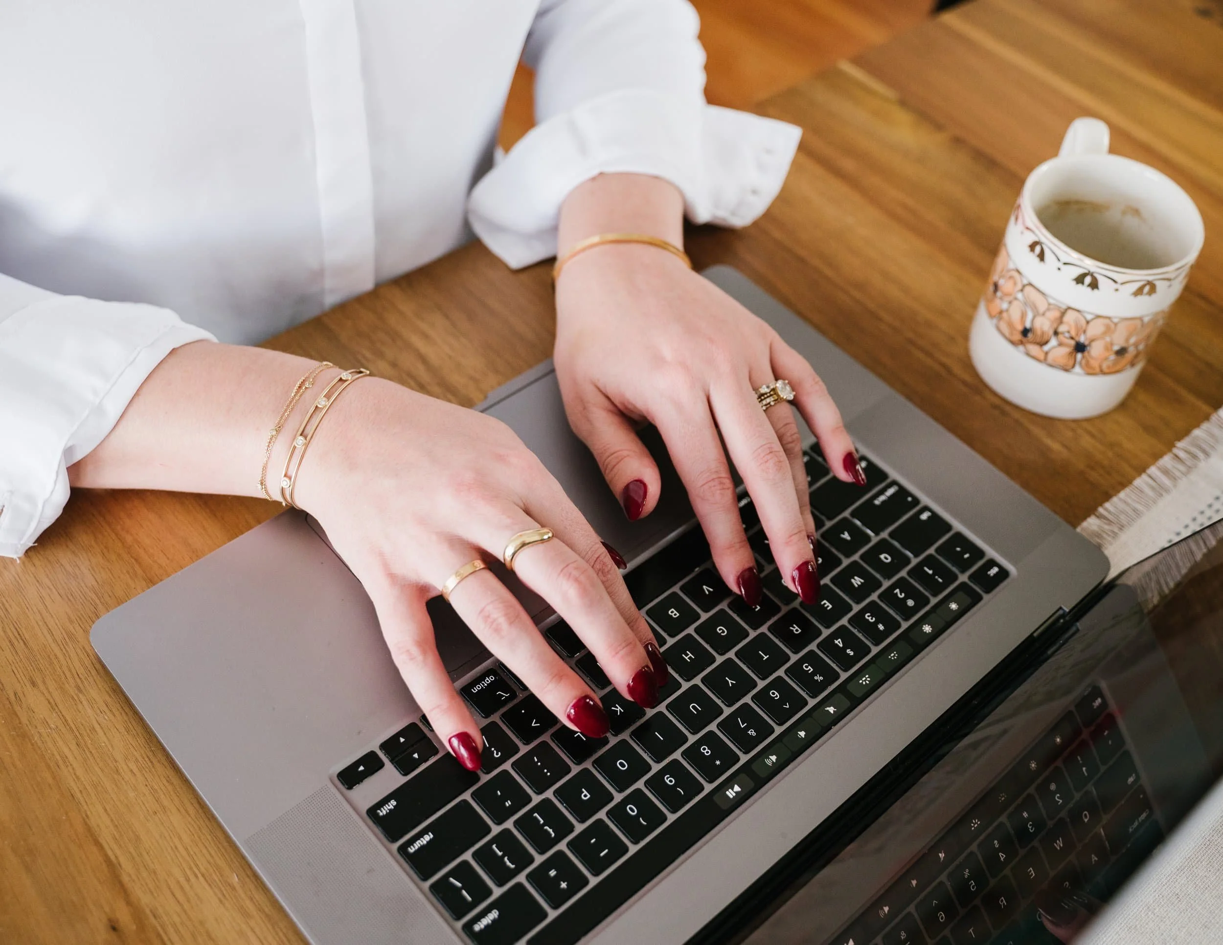 A person with painted red nails typing on a silver laptop keyboard. They're wearing multiple gold rings and bracelets. There is a floral-patterned mug with a beverage and a white and beige cloth placed on a wooden table.