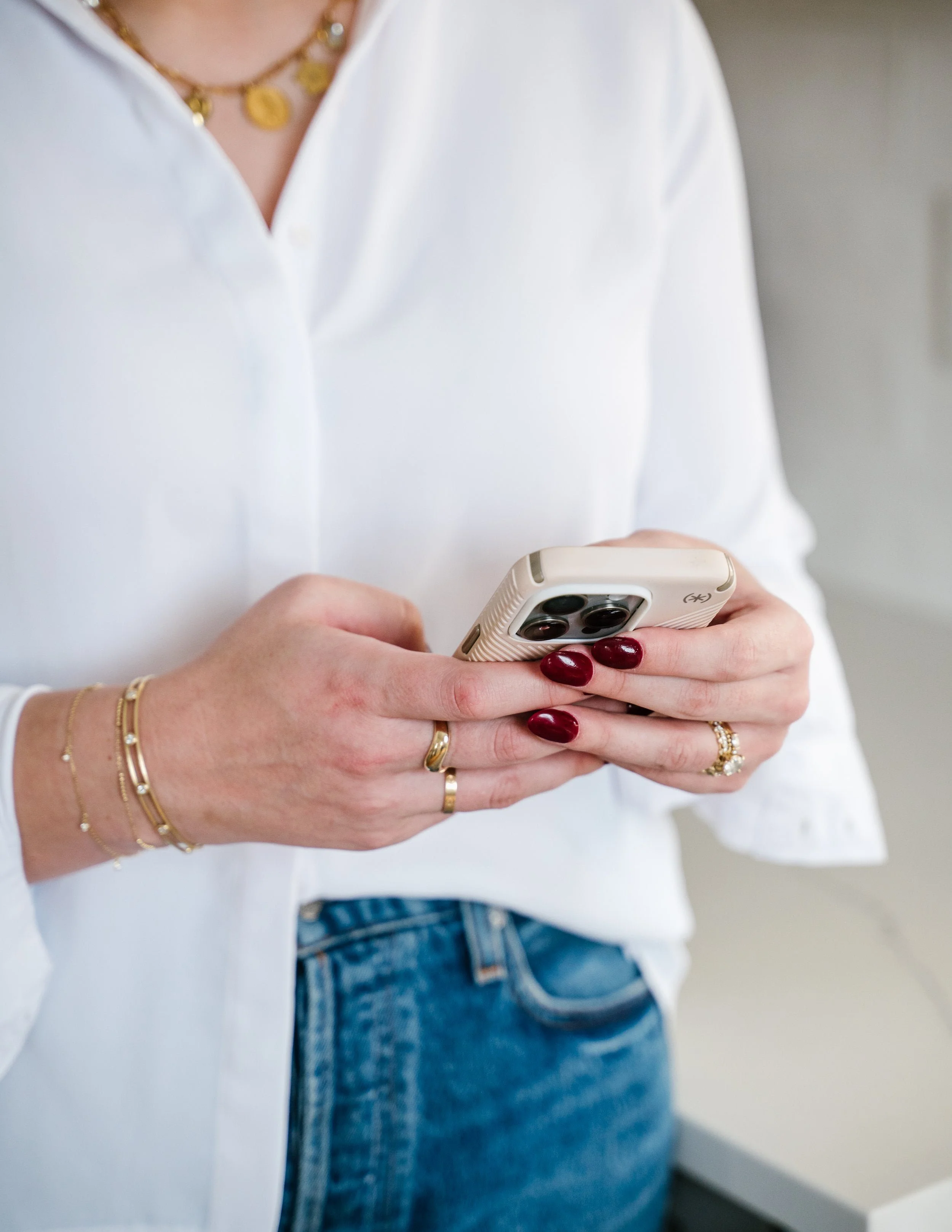 Person wearing a white shirt and blue jeans, holding a smartphone with both hands, displaying multiple gold rings and red nail polish on fingers, and wearing gold jewelry including necklaces and bracelets.