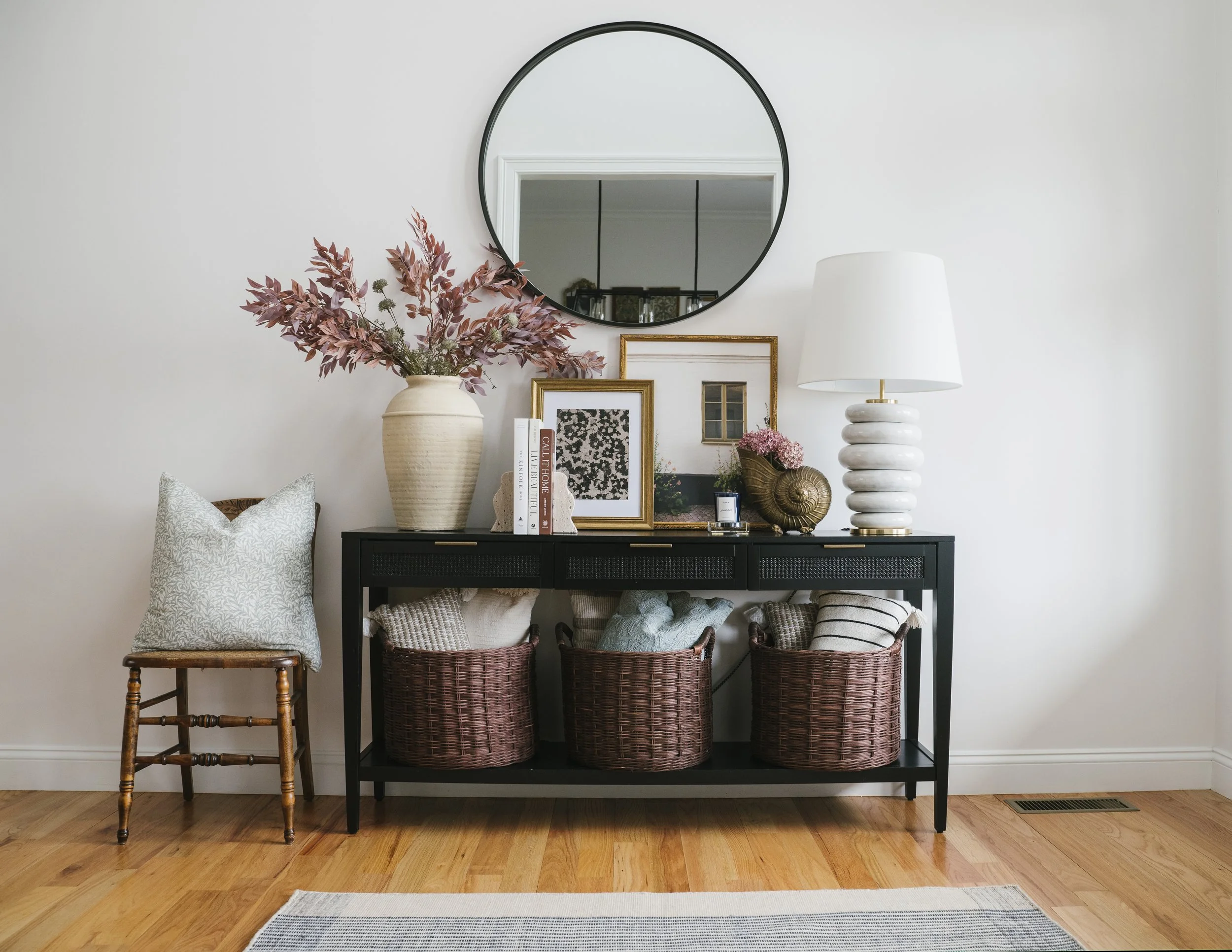 Decorative sideboard against a white wall with framed artwork, a large vase with pink and green foliage, a table lamp, and a mirror above it. The sideboard has three woven baskets with textiles inside, and a small wooden stool with a patterned pillow.
