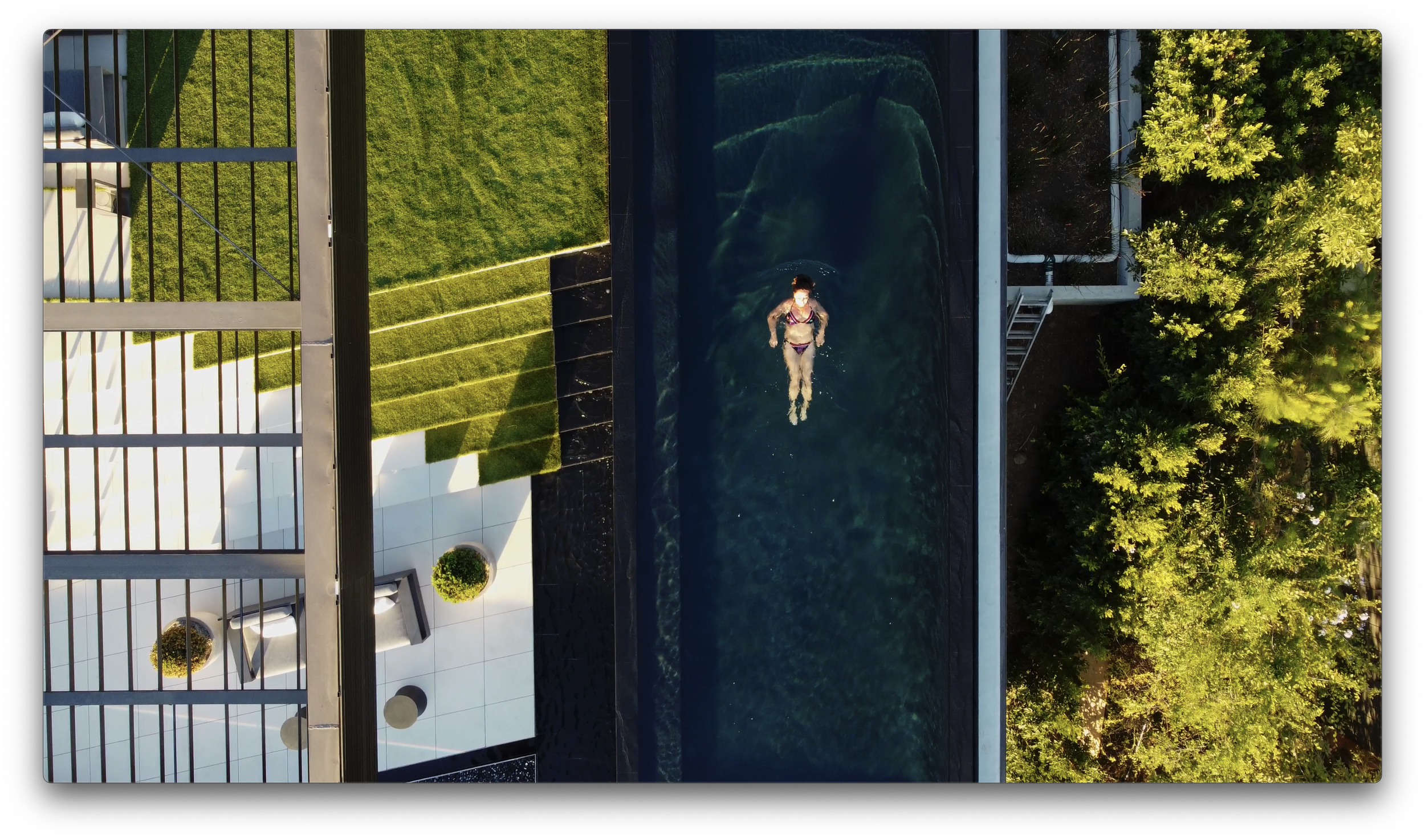 A woman swimming in a dark outdoor pool during daytime, with a garden area and trees around the pool.