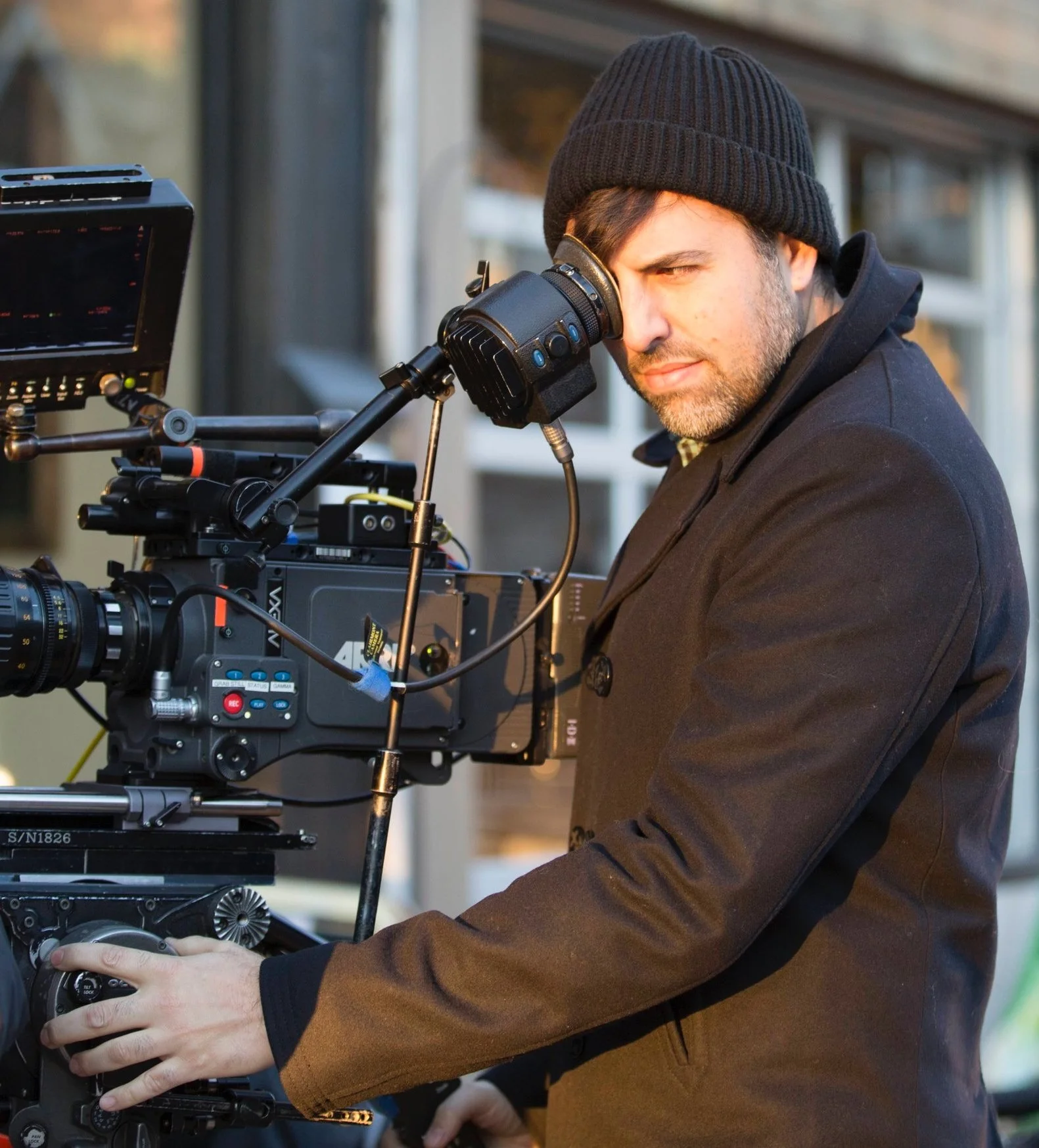 Man wearing a black beanie and dark jacket operating professional film equipment outdoors.