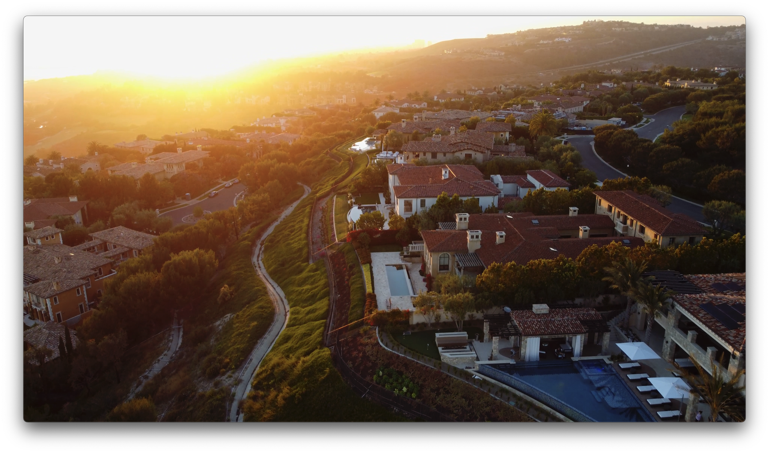 An aerial view of a residential neighborhood at sunset, showing houses with red-tiled roofs, winding streets, and lush green trees and bushes.