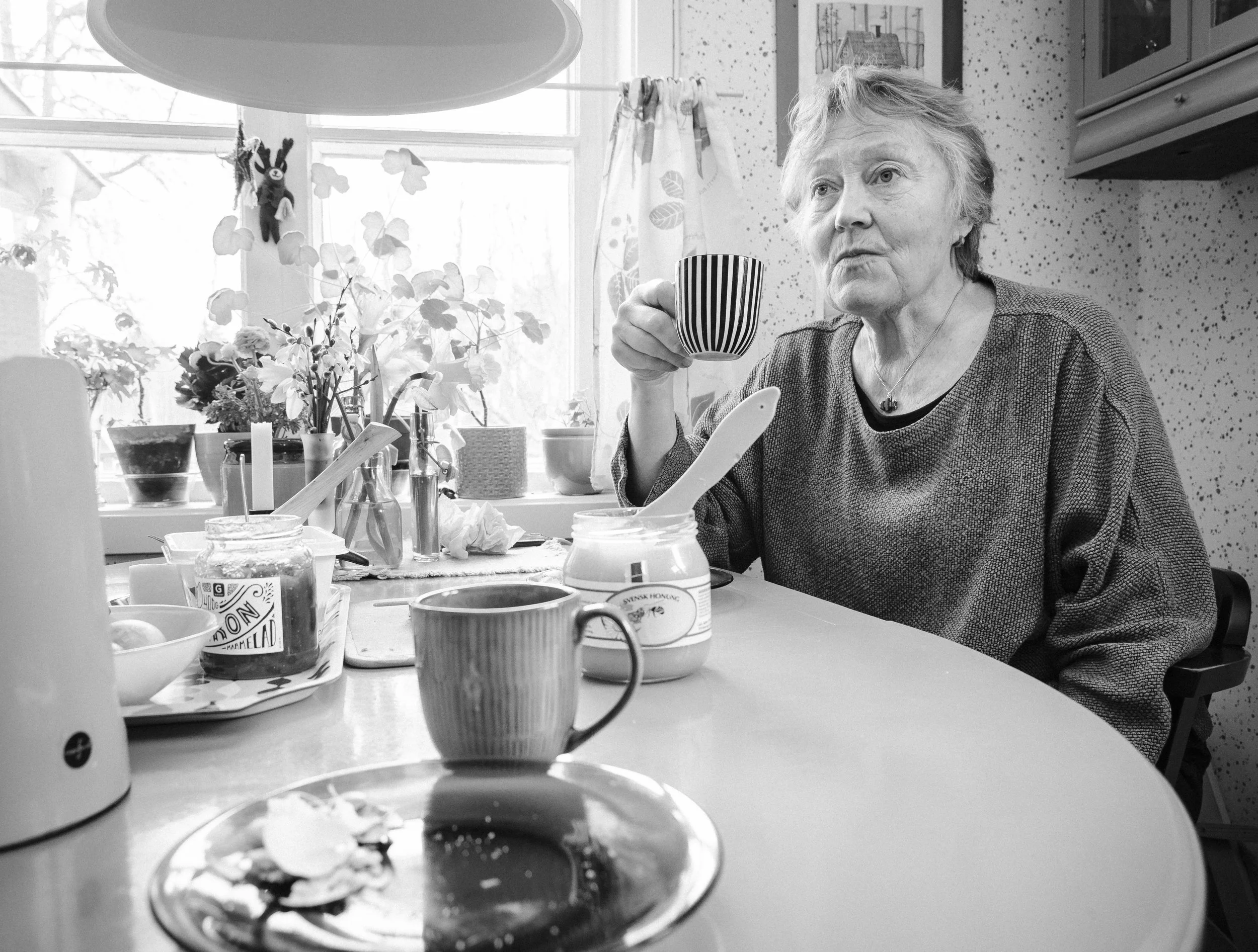 En äldre kvinna sitter vid ett köksbord, håller en mugg med en svart och vit randig design, och ser fundersam ut. Det finns påläggssmör, en kopp, och en burk med marmelad på bordet, samt blommor och köksskåp i bakgrunden.