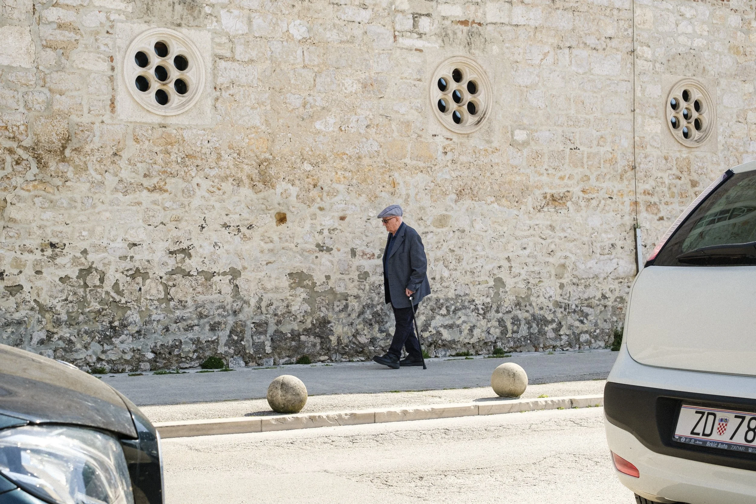 En äldre man i mörkblå jacka och keps går med en käpp längs en gata, i bakgrunden en gammal tegelvägg.
