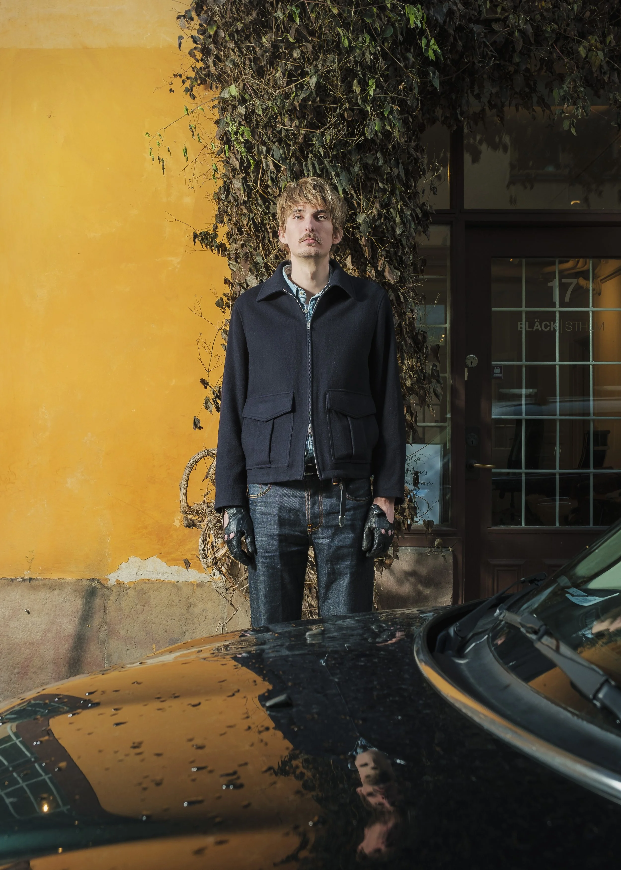 En man står utomhus framför en vägg täckt av murgröna, med en gul vägg i bakgrunden. Han är klädd i en svart jacka och jeans, och bär handskar. En svart bil reflekteras i fronten av bilen framför honom.