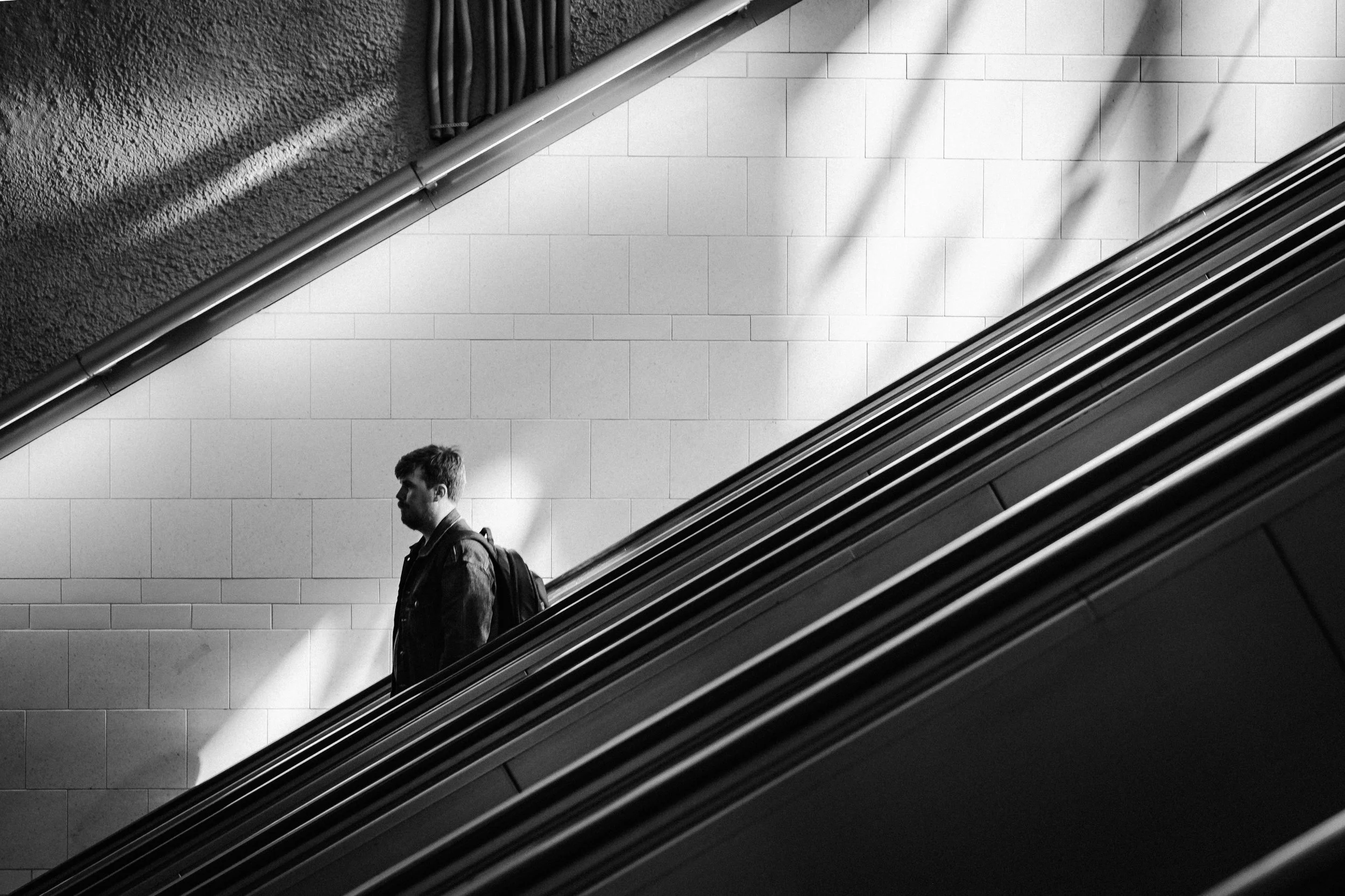 En silhuett av en man med ryggsäck på en rulltrappa i ett svartvitt foto, med en vit tegelvägg i bakgrunden.