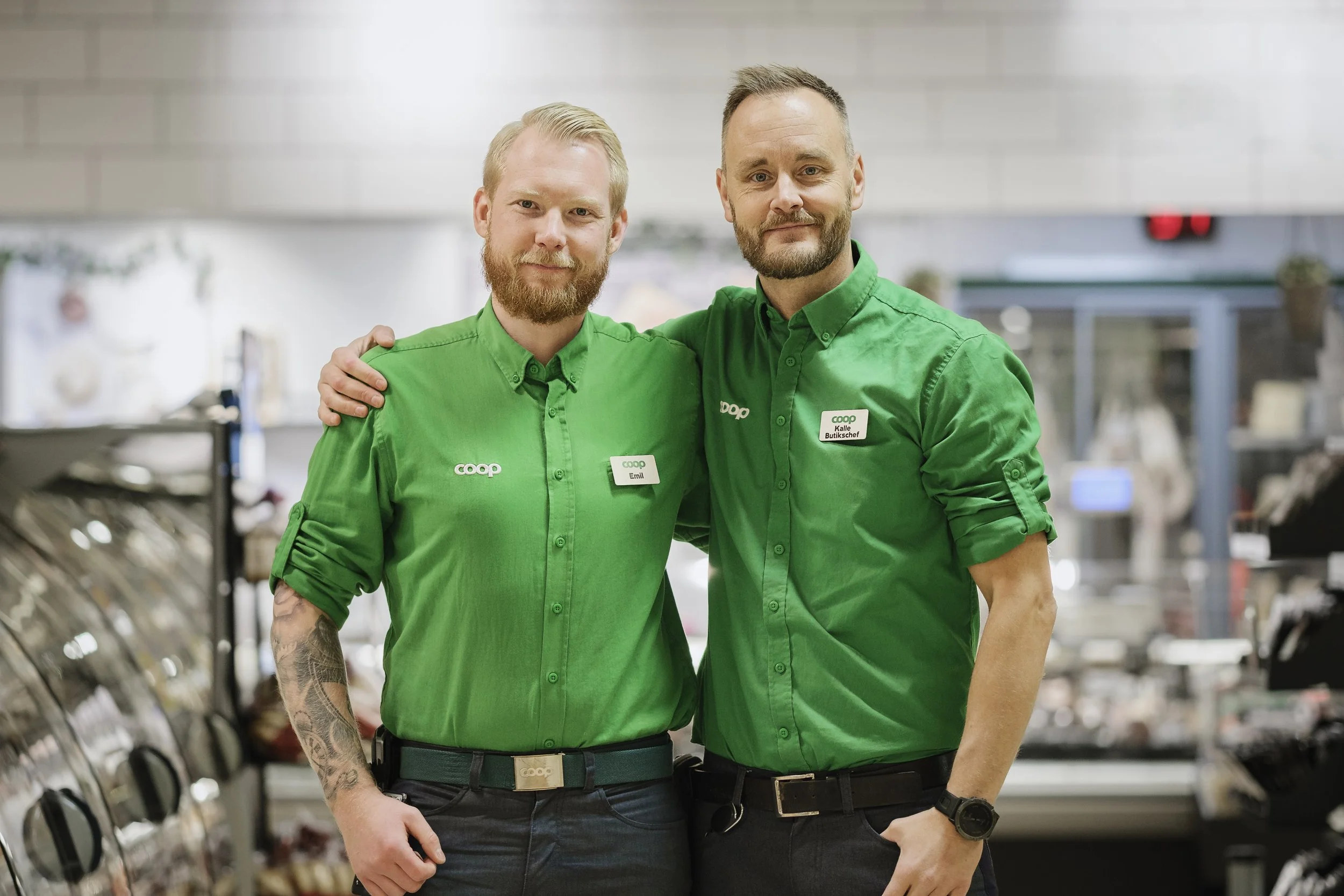 Två män i gröna arbetsenda kläder som står i en butik och ler mot kameran, en av dem har tatuering på armen.