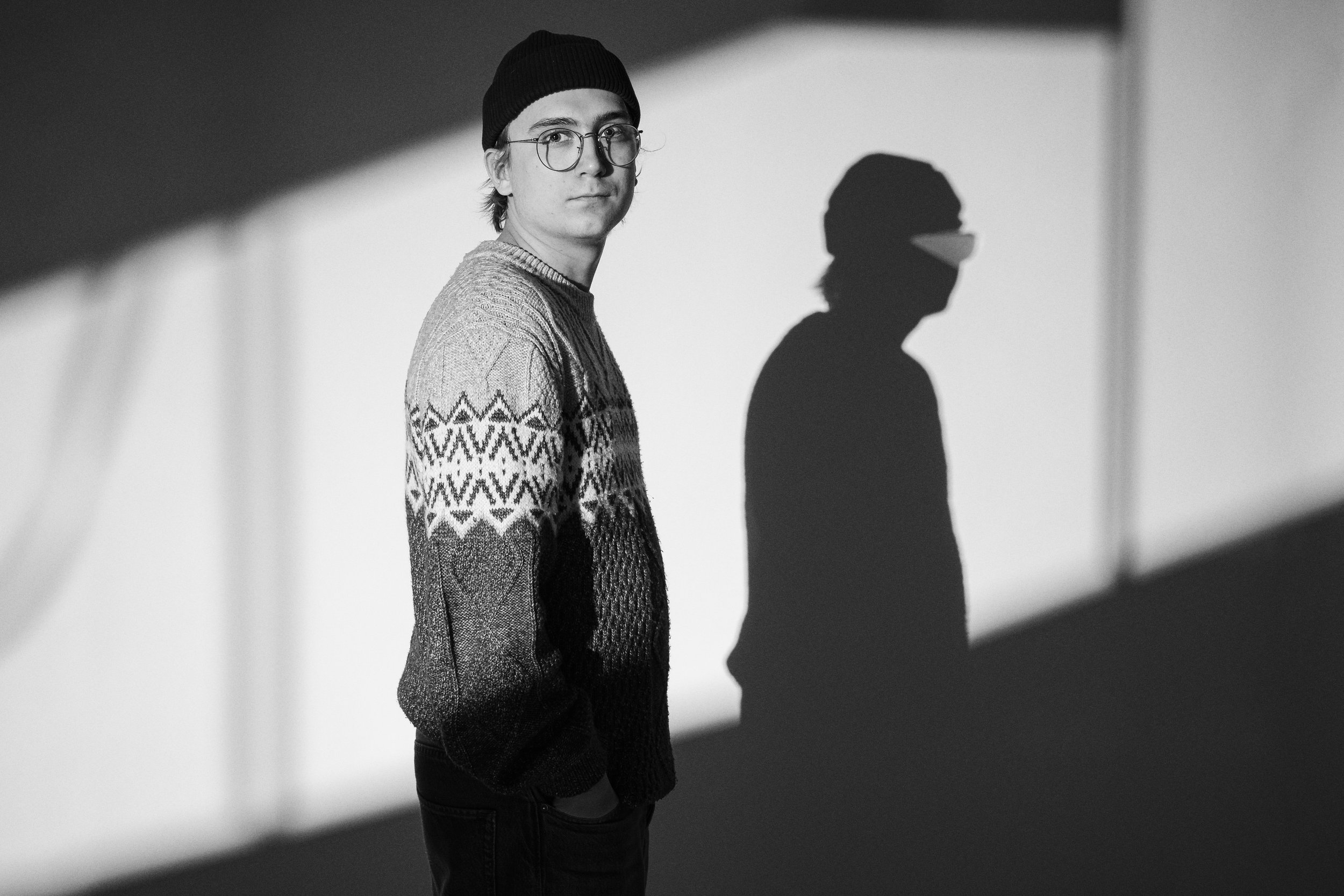 En ung man med glasögon, mössa och en tröja med mönster, står i ett rum med skuggor på väggen.