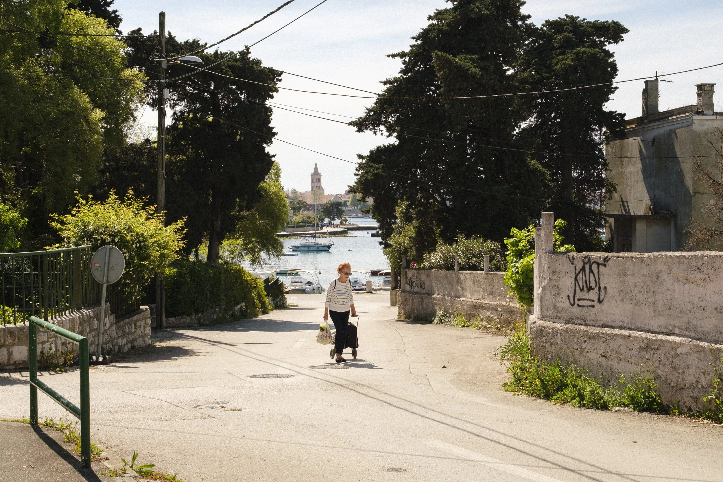 En äldre kvinna går på en gata med väska och shoppingkassar, precis vid vattnet med båtar och en kyrktorn i bakgrunden.