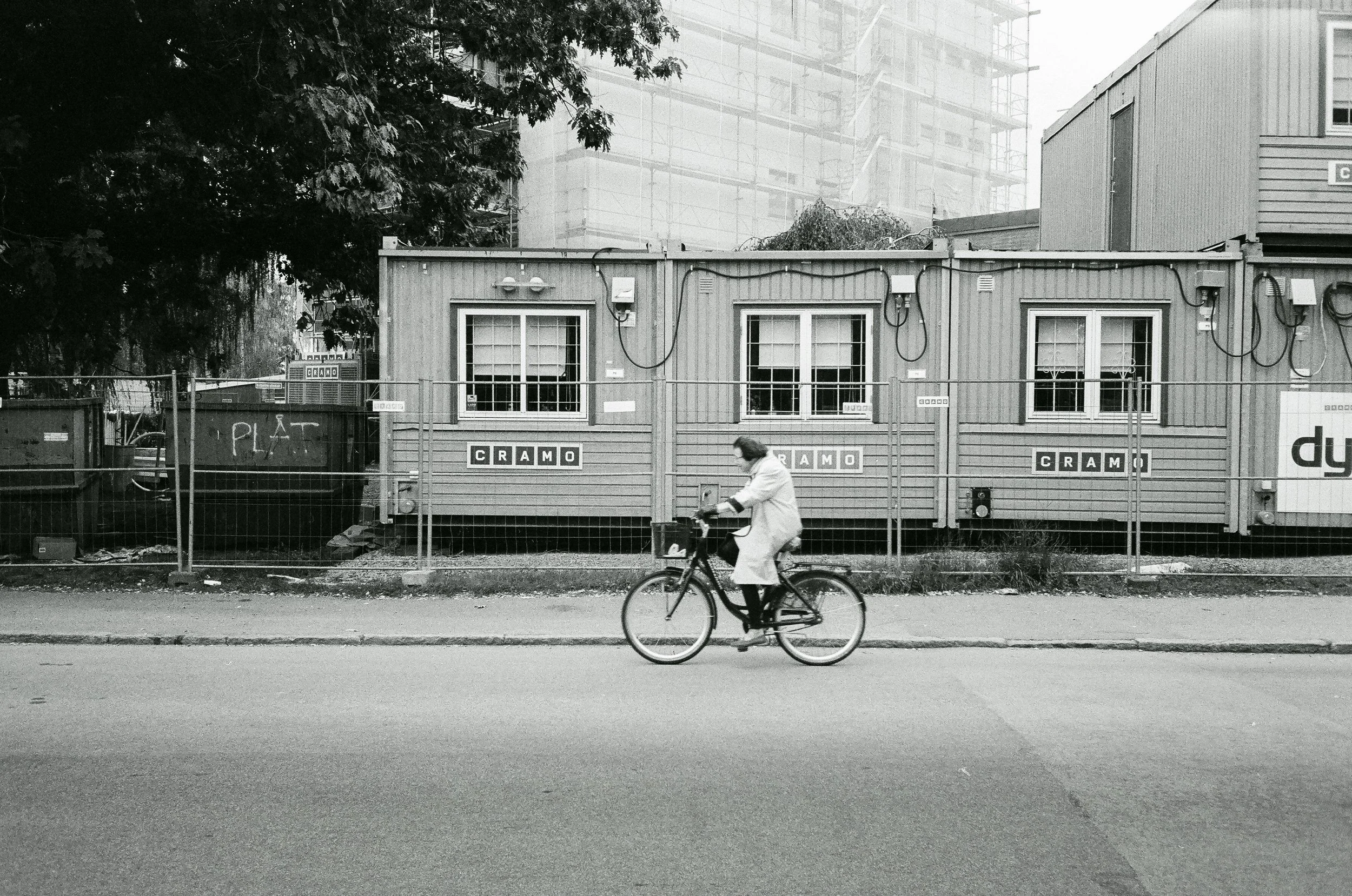 En person som cyklar på en gata framför en byggnad med tre fönster och en skylt med texten 'CRAMO'.