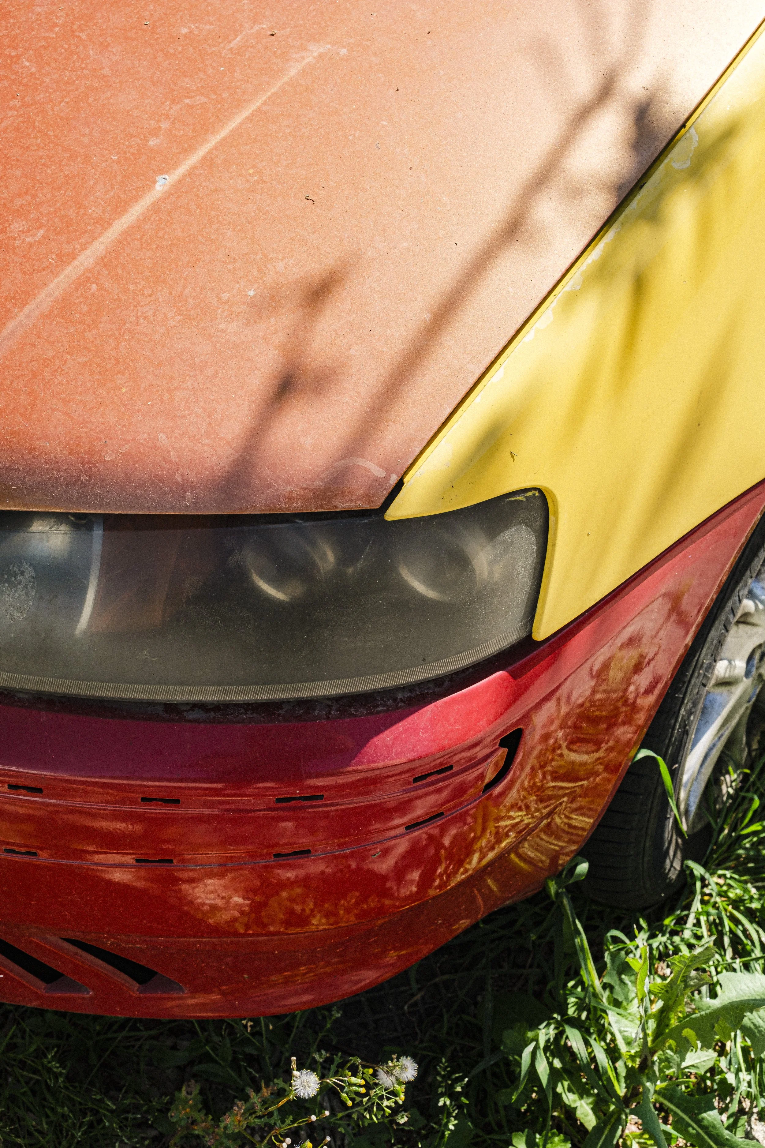 Tvärställd bild av ett bilhuvud med en förfallen och smutsig bil i rött och gult färgschema, omgiven av gräs och växter.