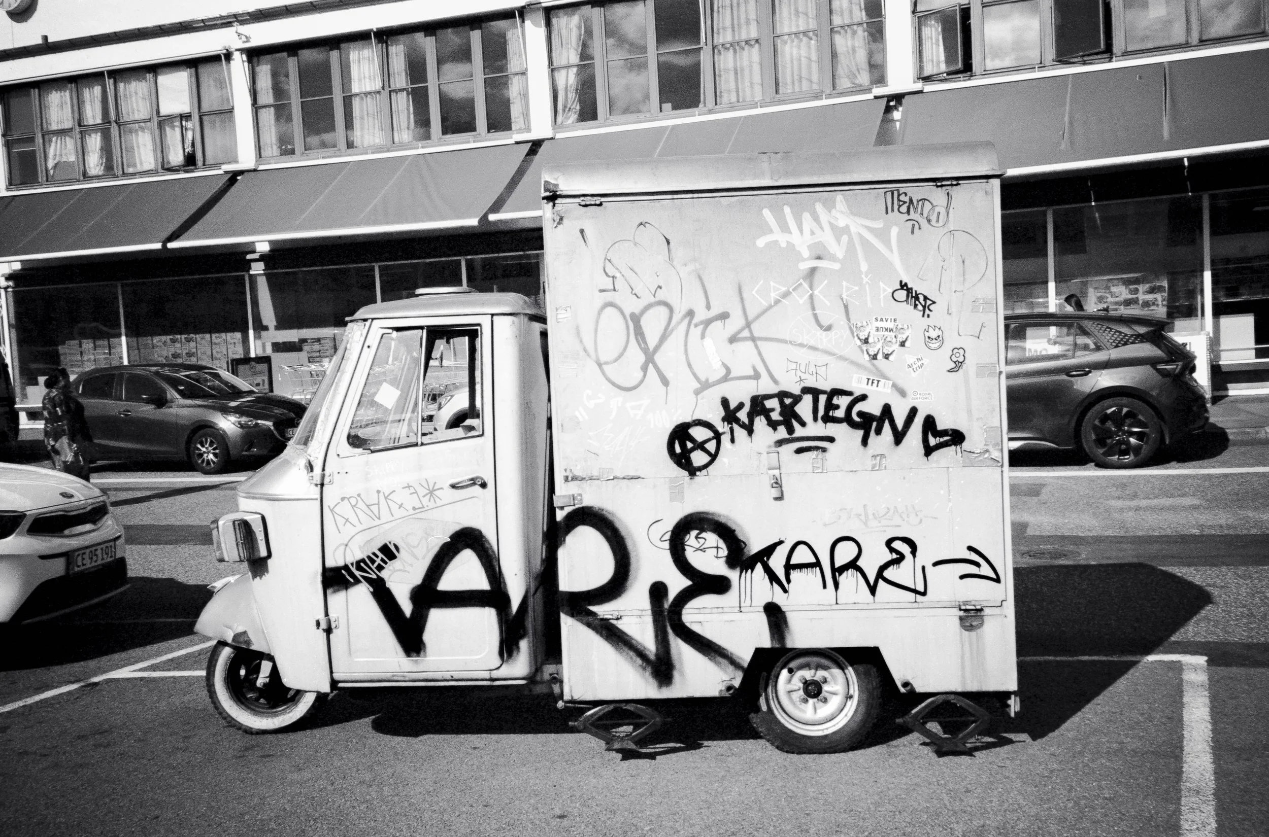 En graffiti-dekorerad liten vit trilskona fordon på en gata, med parkerade bilar och en butik i bakgrunden.