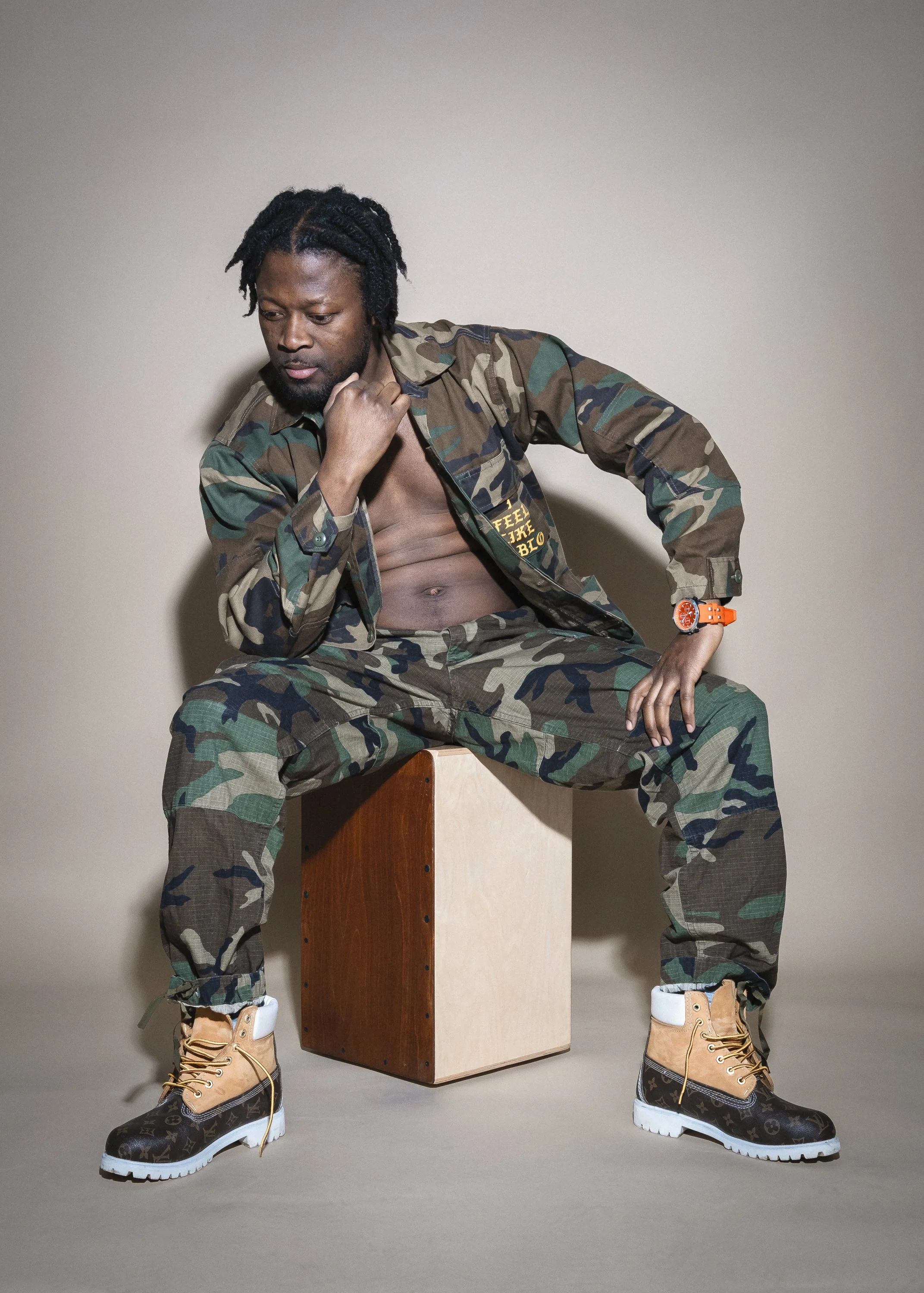 En man i camouflagekläder som sitter på en träbox, med ett skärp och stövlar från Louis Vuitton, mot en neutral bakgrund.