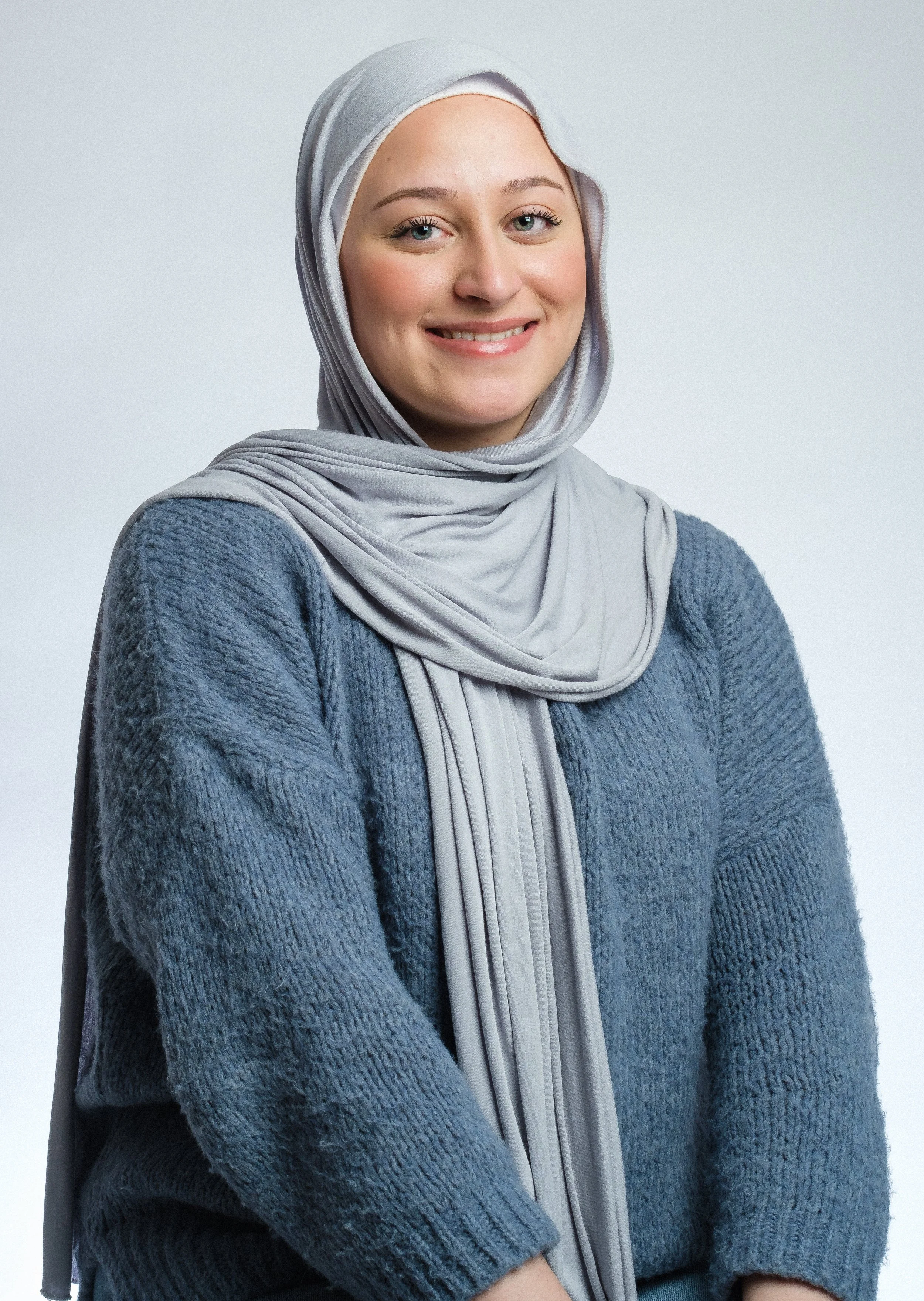 en ung kvinna bär en ljusgrå slöja och en blå stickad tröja, ler mot kameran