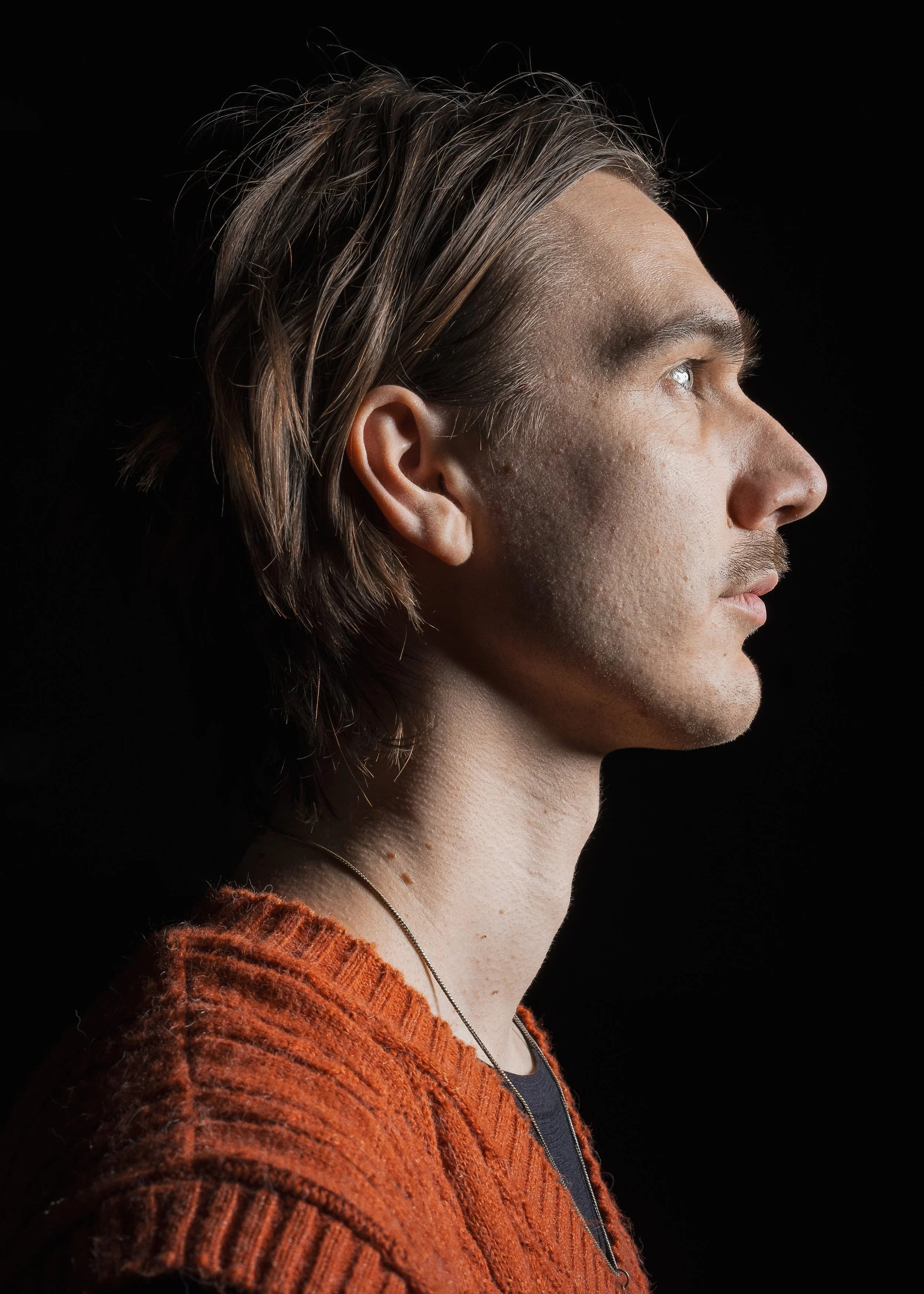 Porträtt av en man i profil mot svart bakgrund, med långt trött hår och kedja runt halsen, bär en orange stickad tröja.