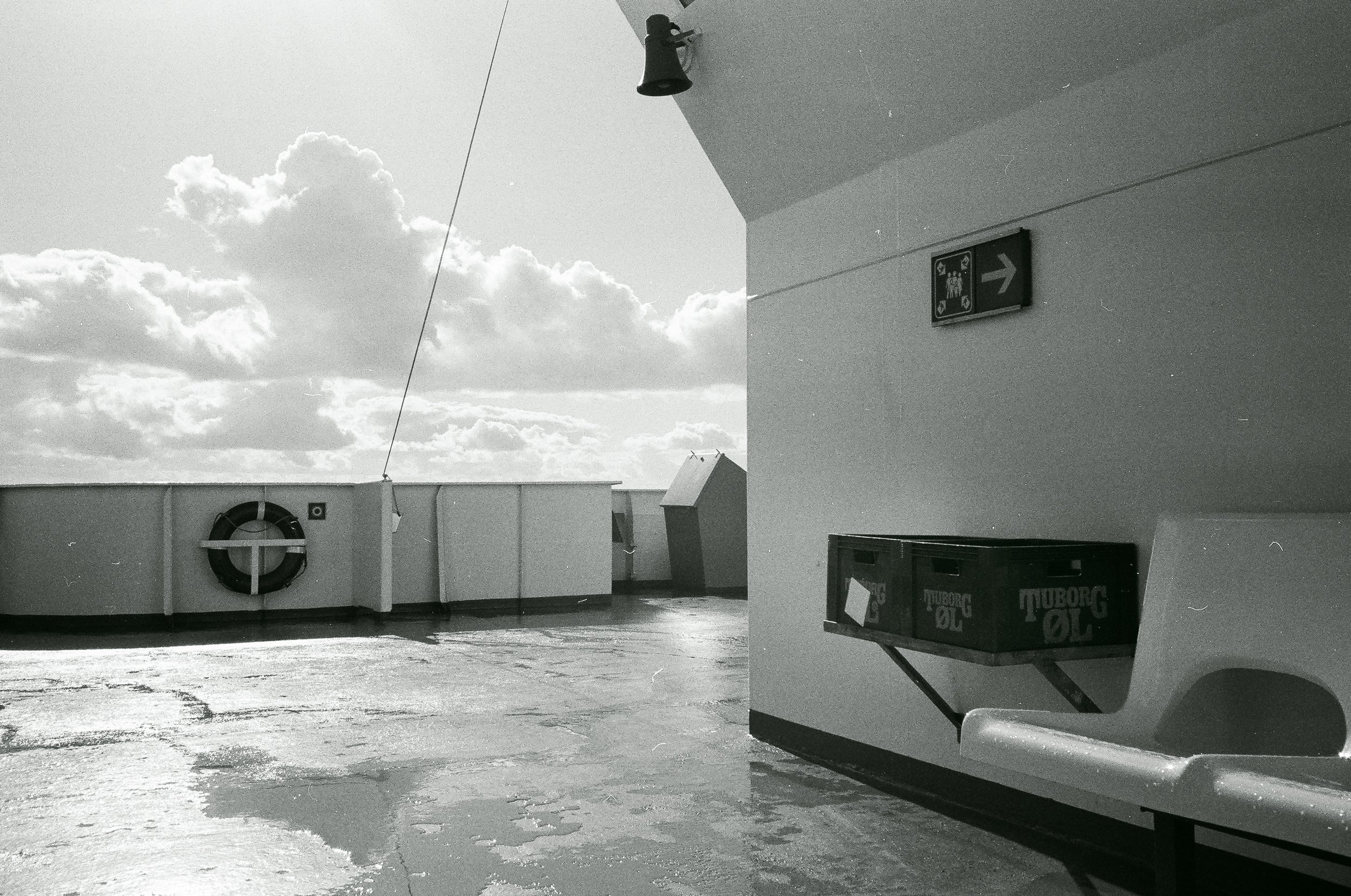 Den svarta och vita bild visar ett tak med en vattentank, ett räddningsringhjul, ett litet hus, en vägg med en skylt och ett rör med ölbackar.