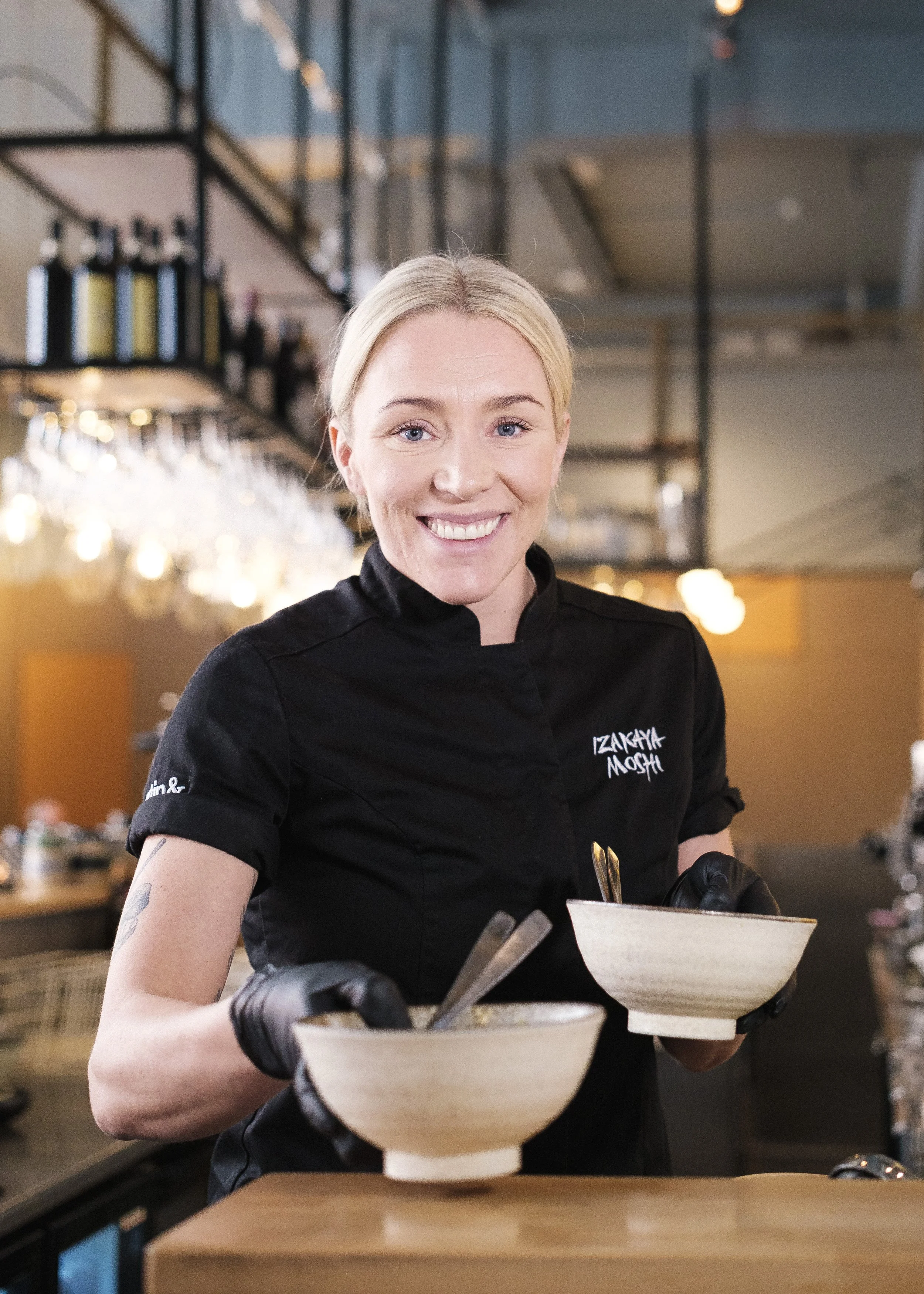 En glad kvinnlig kock i svart uniform med texten 'Izakaya Mough' ler mot kameran i en modern restaurangmiljö och håller i två skålar.