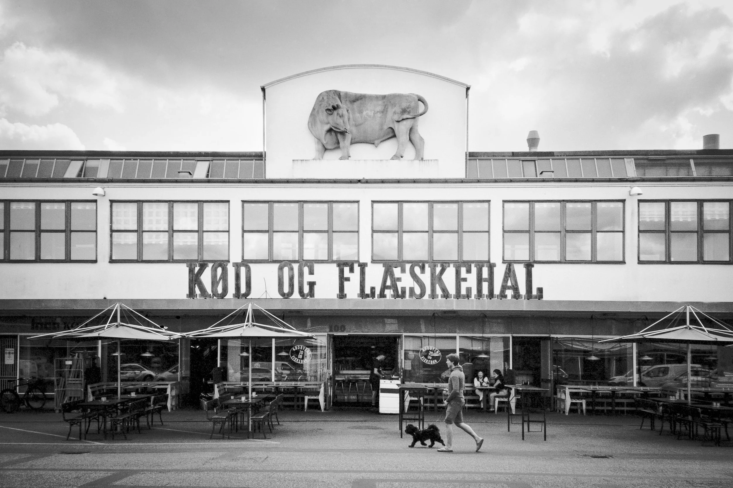 En svartvit bild av ett restaurangutrymme med gågator framför. Restaurangen har ett skyltfönster och uteplats med stolar och bord. På taket finns en stor, tre-dimensionell elefantstaty och texten 'KOD OG FLØSKEHALL'.