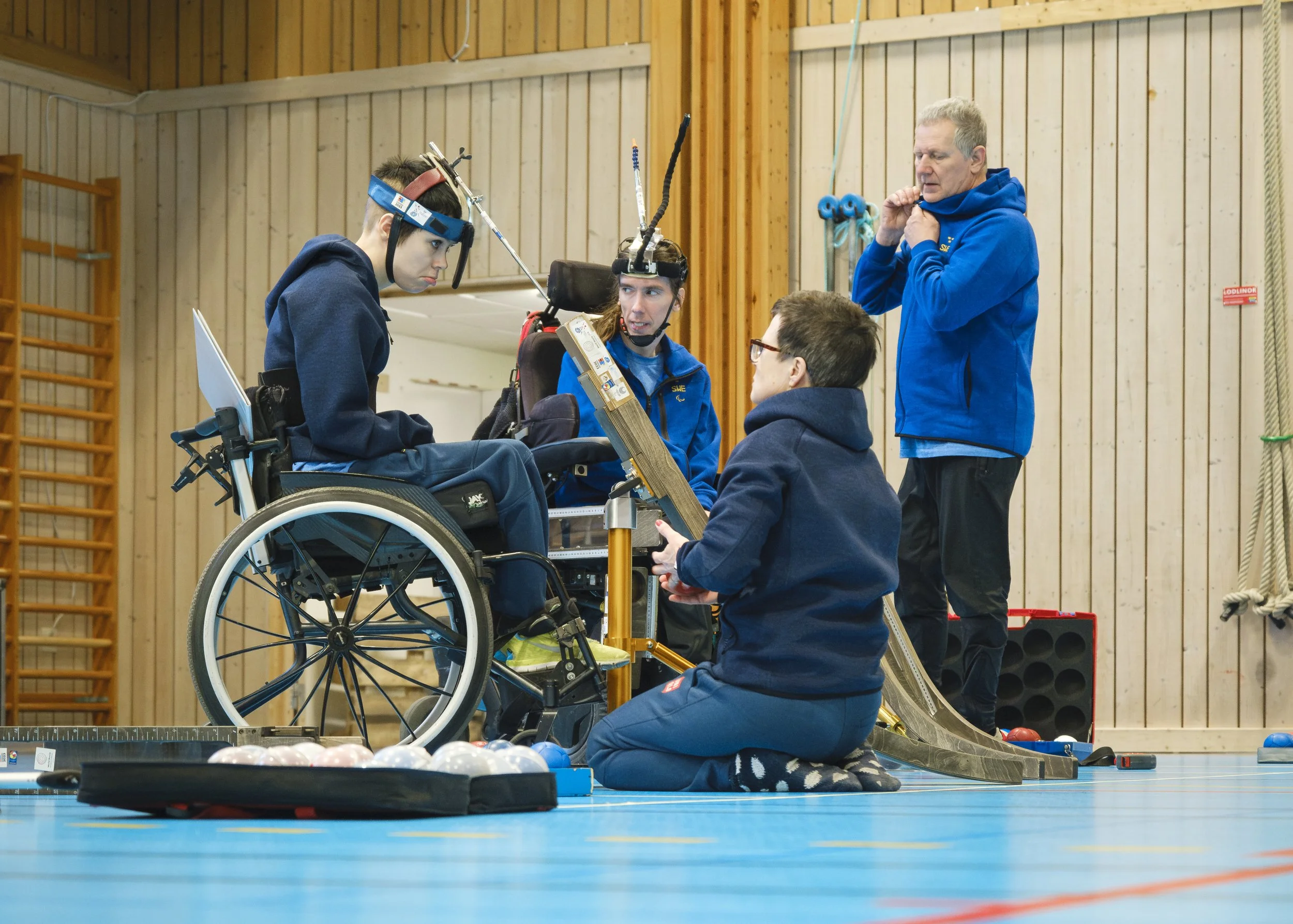 En person i rullstol reciterar bowlingregler för tre andra personer, en av dem sitter på golvet, medan två andra står bredvid. De alla är i en inomhus sporthall, och det finns bowlingbollar på golvet.