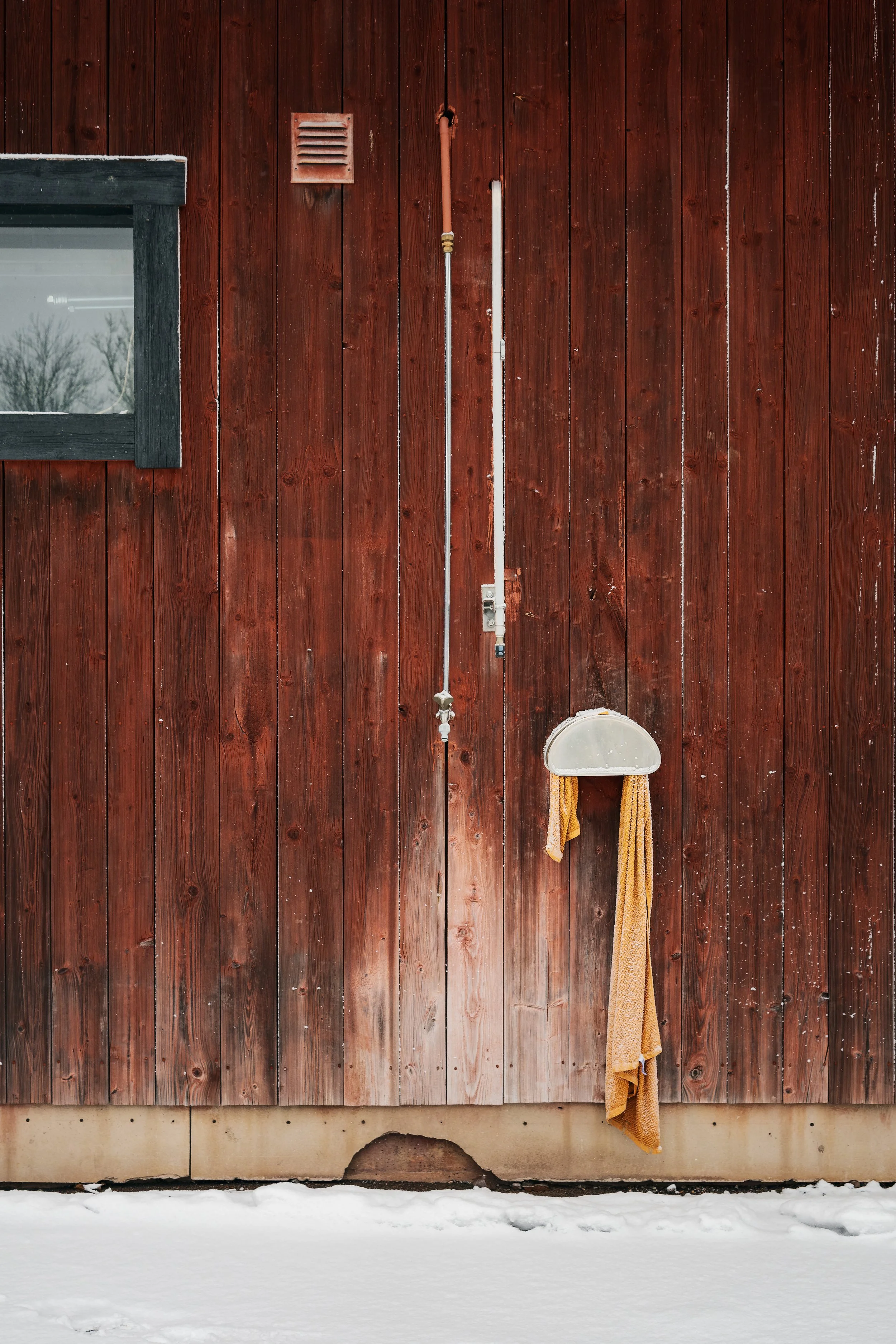 Röd trävägg med ett fönster, ett ventilationsgaller, ett rör, en hängande borste och en liten skylt.