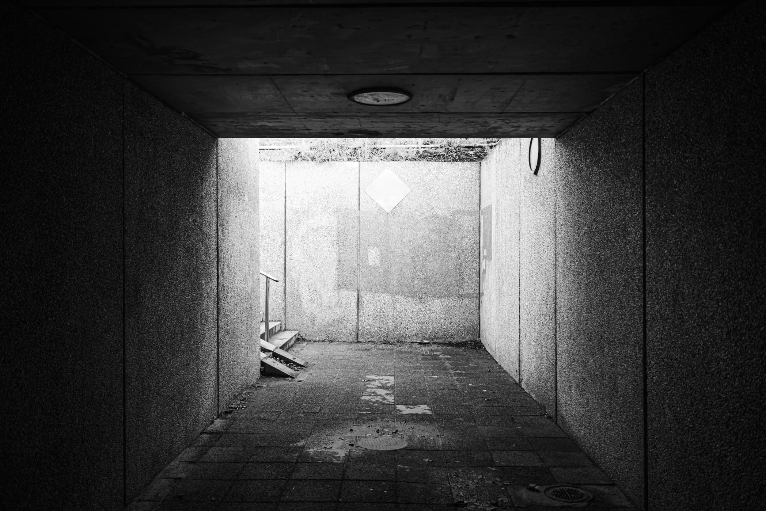 En svartvit bild av en tunnel under ett byggnad med väggar av betong och ett golv av plattor, som leder ut till en ljusare plats utomhus.