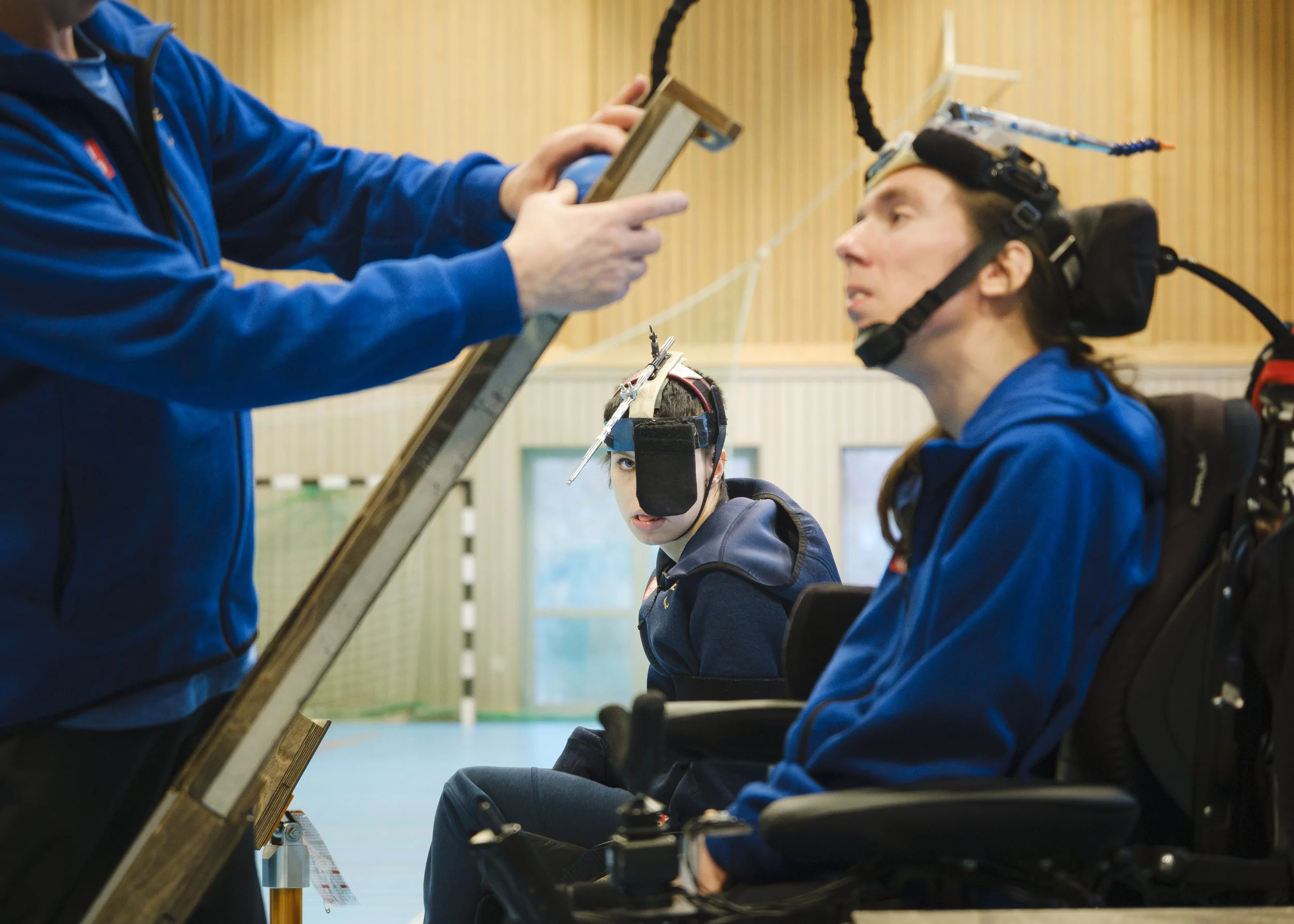 Två personer med hjälpmedel för bedömning av sinnesfunktion i ett gymnastikhall, en sitter i rullstol och den andra har en elektrodiagnostisk utrustning på huvudet. En person håller i en mätare eller kontrollinstrument.