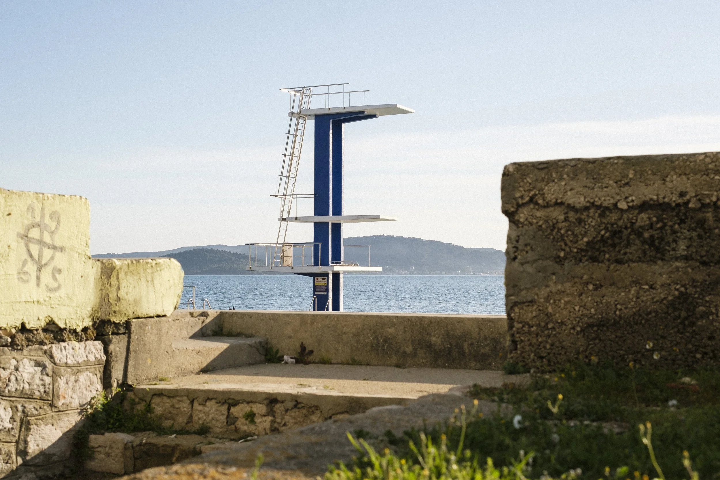 En översiktsbild av en simklubb på en strand med en hopptrappa i mitten, omgiven av murar och en havsvik i bakgrunden.