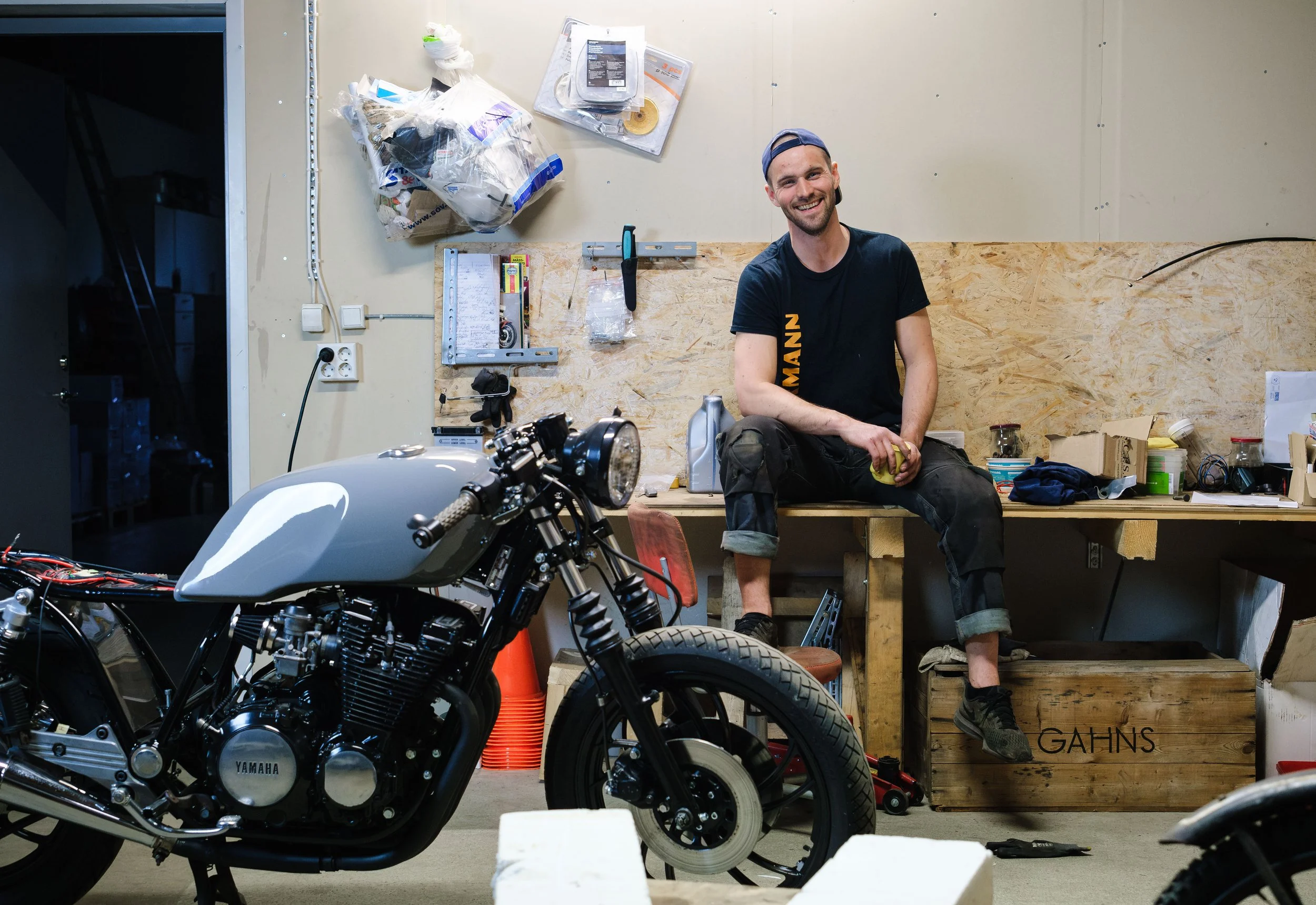 En glad man som sitter på en arbetsbänk i ett verkstadssjukhus bredvid en svart Yamaha-motorcykel. Verkstaden innehåller verktyg, prylar och en trälåda med namnet 'GAHNS'.