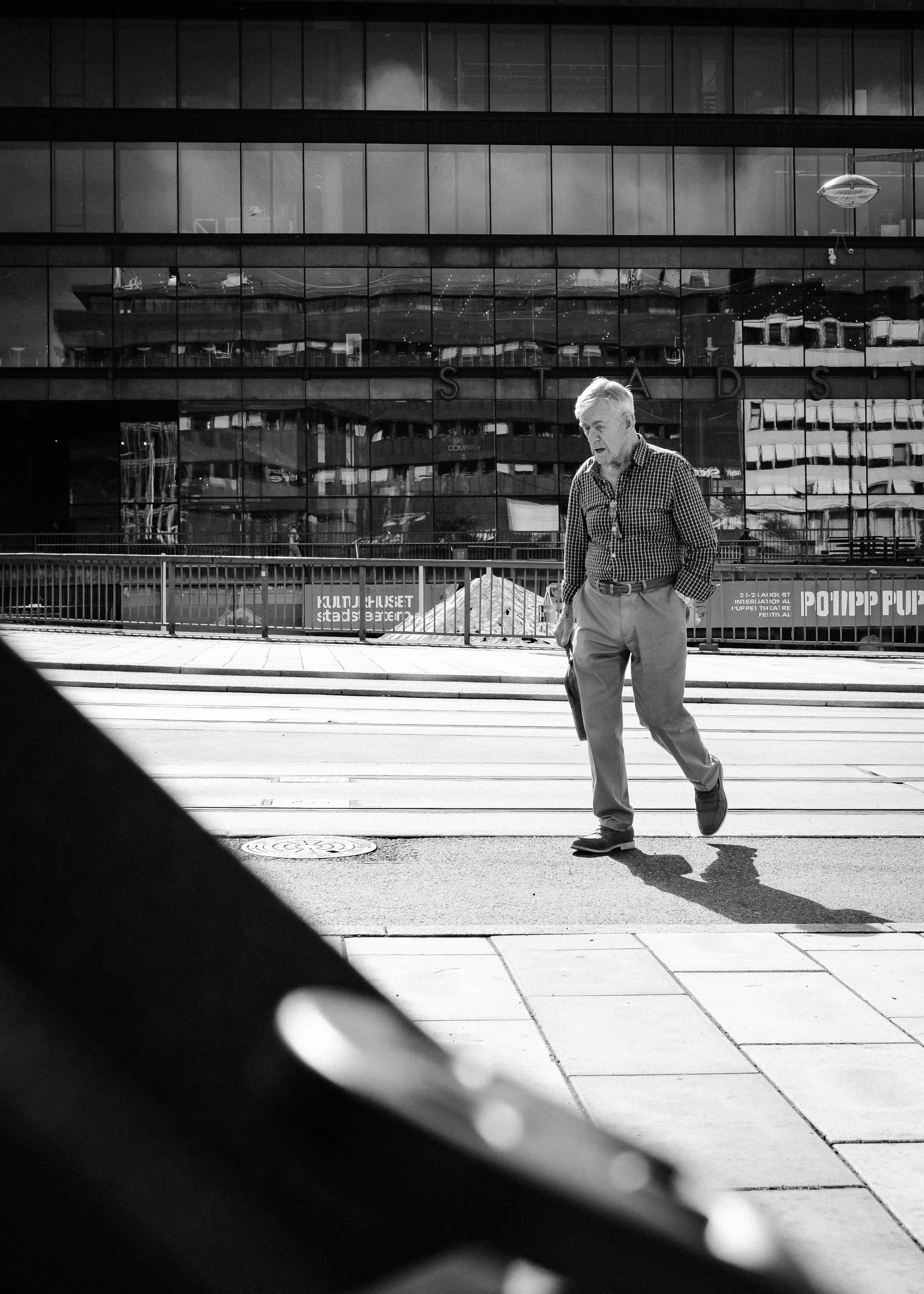 En man går över en stadsgata med moderna byggnader i bakgrunden, syns i svartvitt.