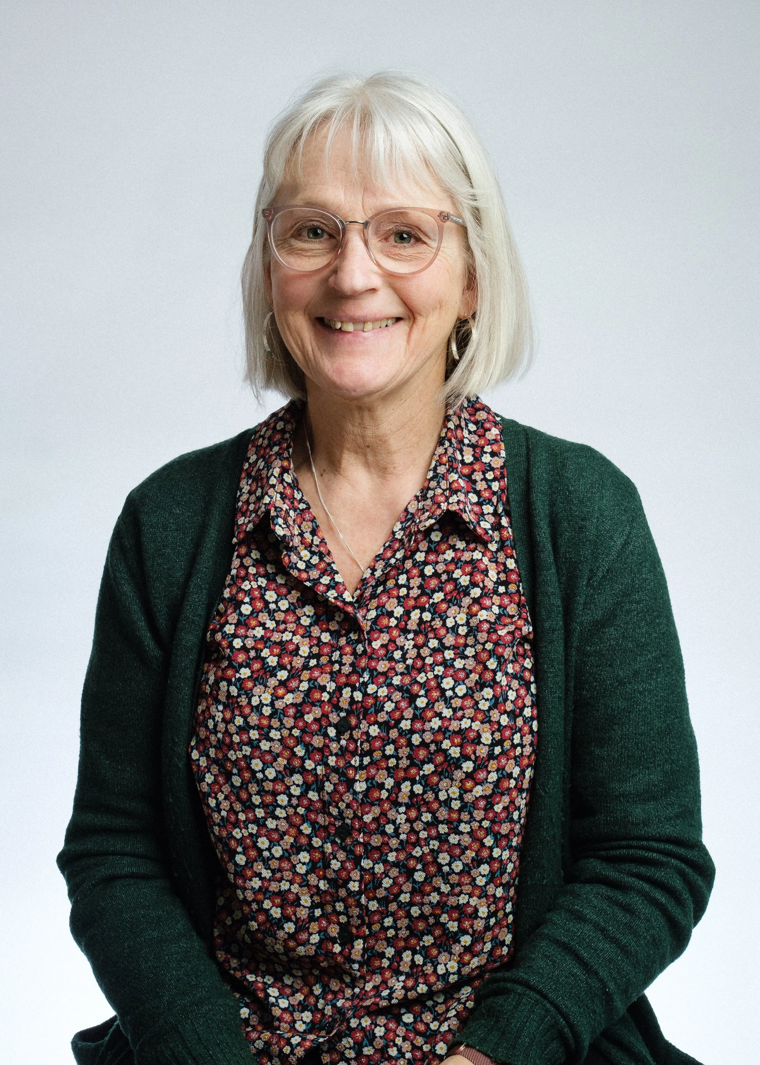 en kvinna med rakt, vitt hår, glasögon och bekväm klädsel, sitter mot en ljus bakgrund