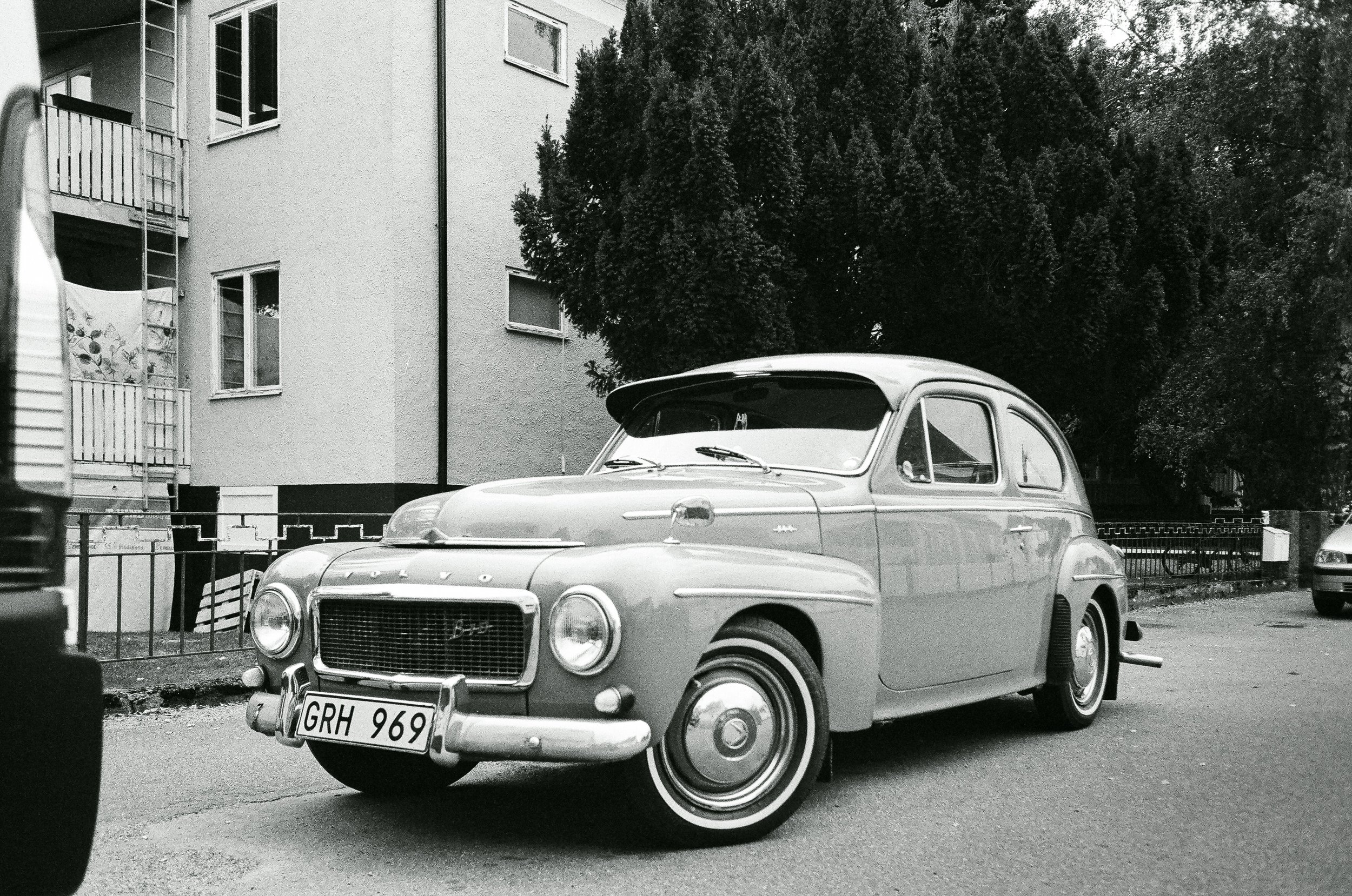 En svartvit bild av en äldre bil parkerad på en gata, med ett flerfamiljshus och träd i bakgrunden.