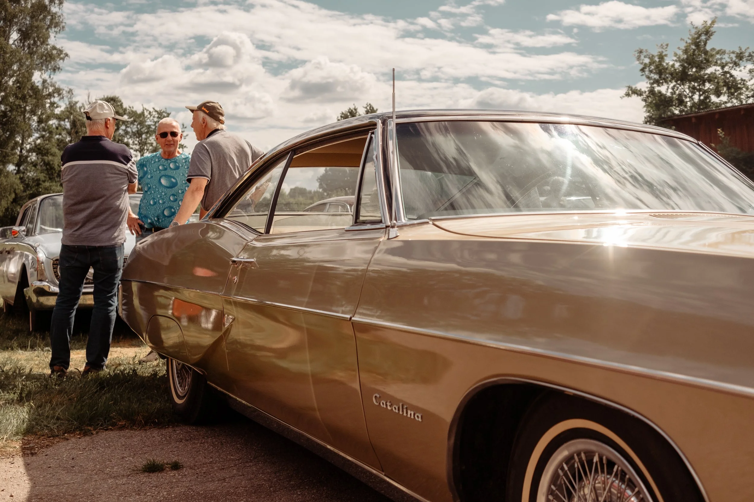 Tre personer som står och pratar bredvid en klassisk gul bil, med en annan bil i bakgrunden, på en gräsplätt under en molnig himmel.