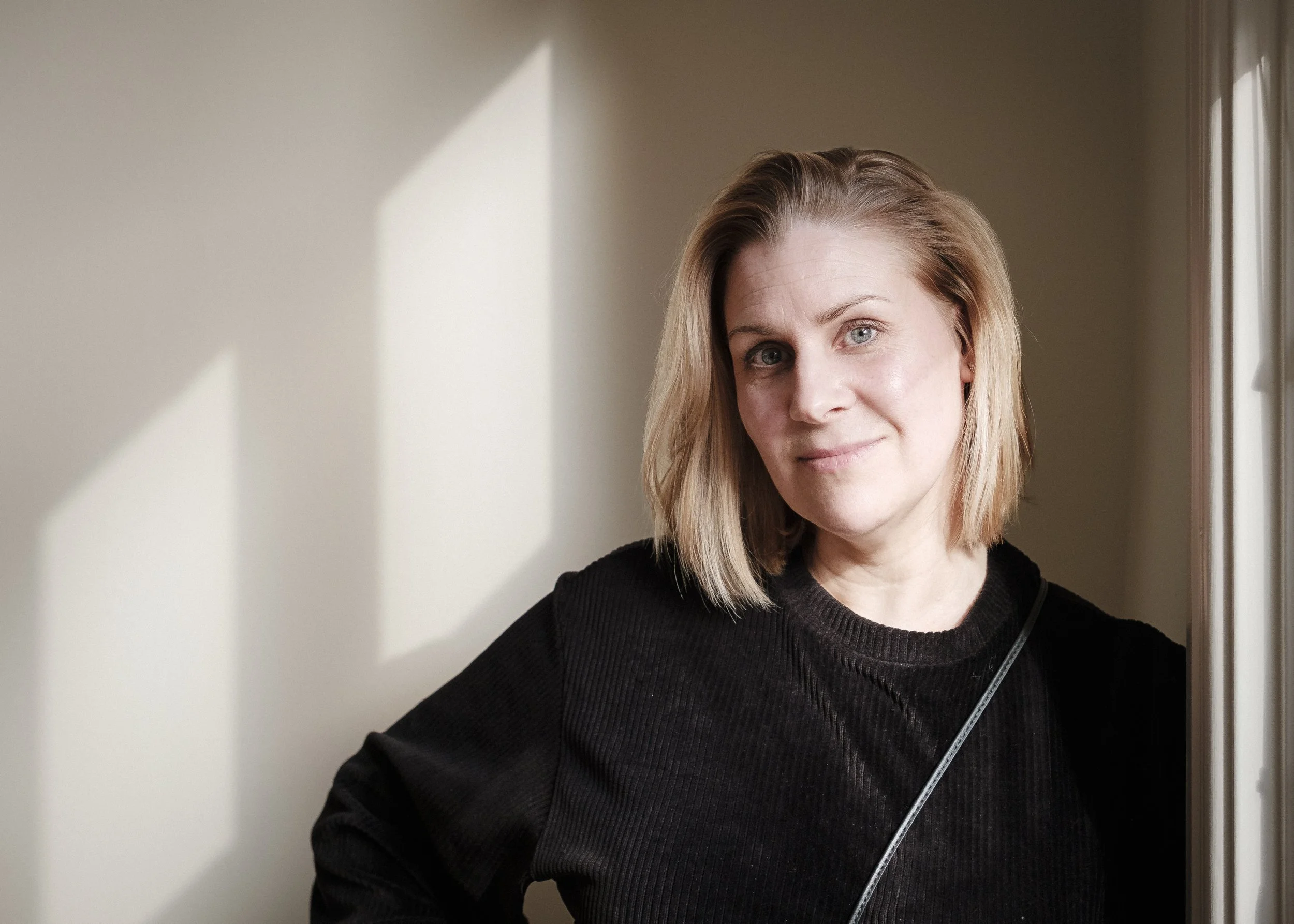 Kvinna med ljusbrunt, axellångt hår, som lutar sig mot en vägg med skuggor som formar kvadratiska mönster, i ett rum med neutrala väggar.